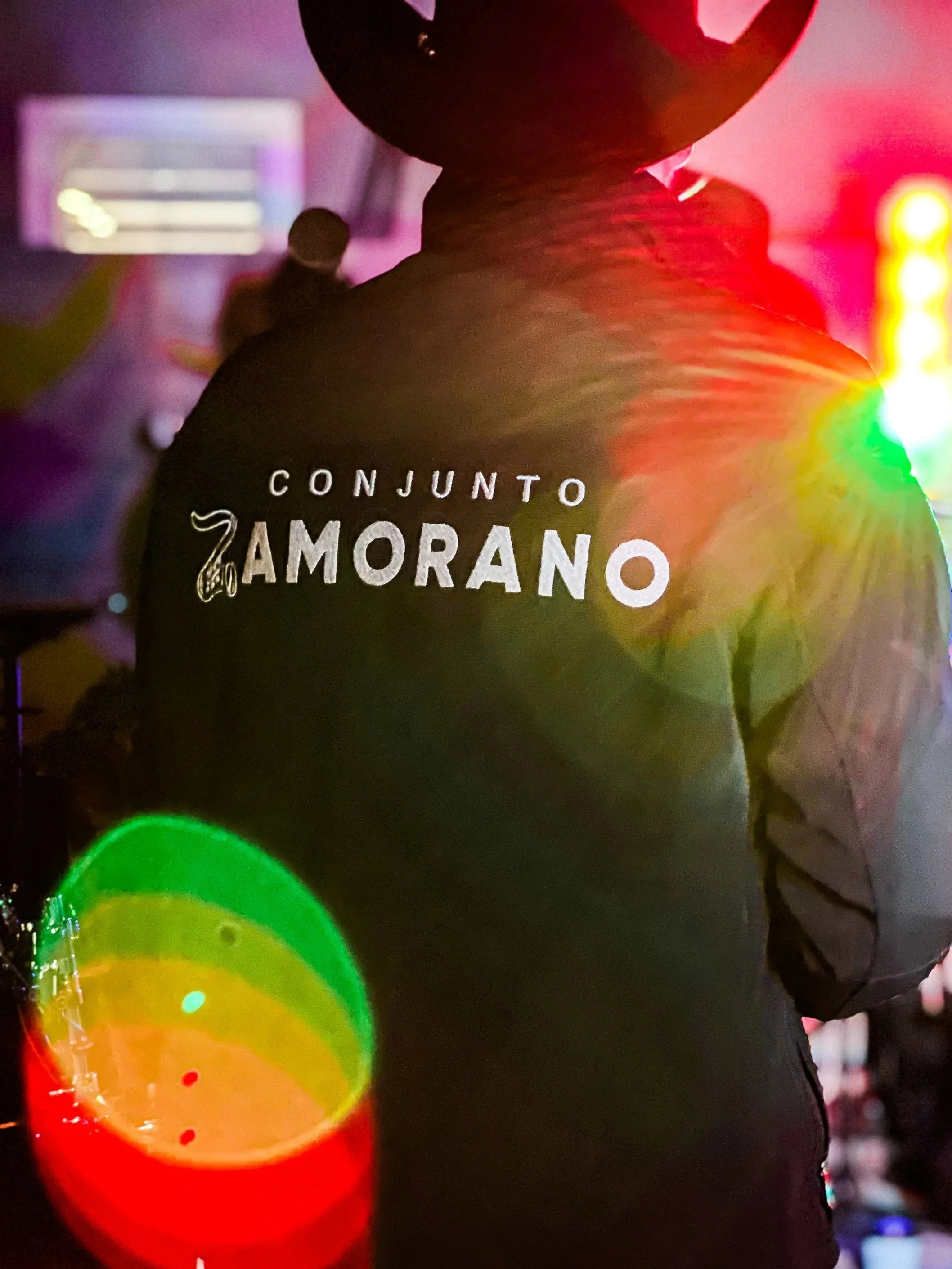 Una persona vista de espaldas, usando una camiseta negra con el texto 'CONJUNTO AMORANO' y un dibujo de una serpiente. En el fondo, luces de colores y un ambiente oscuro, con un vaso con luces de colores en primer plano.