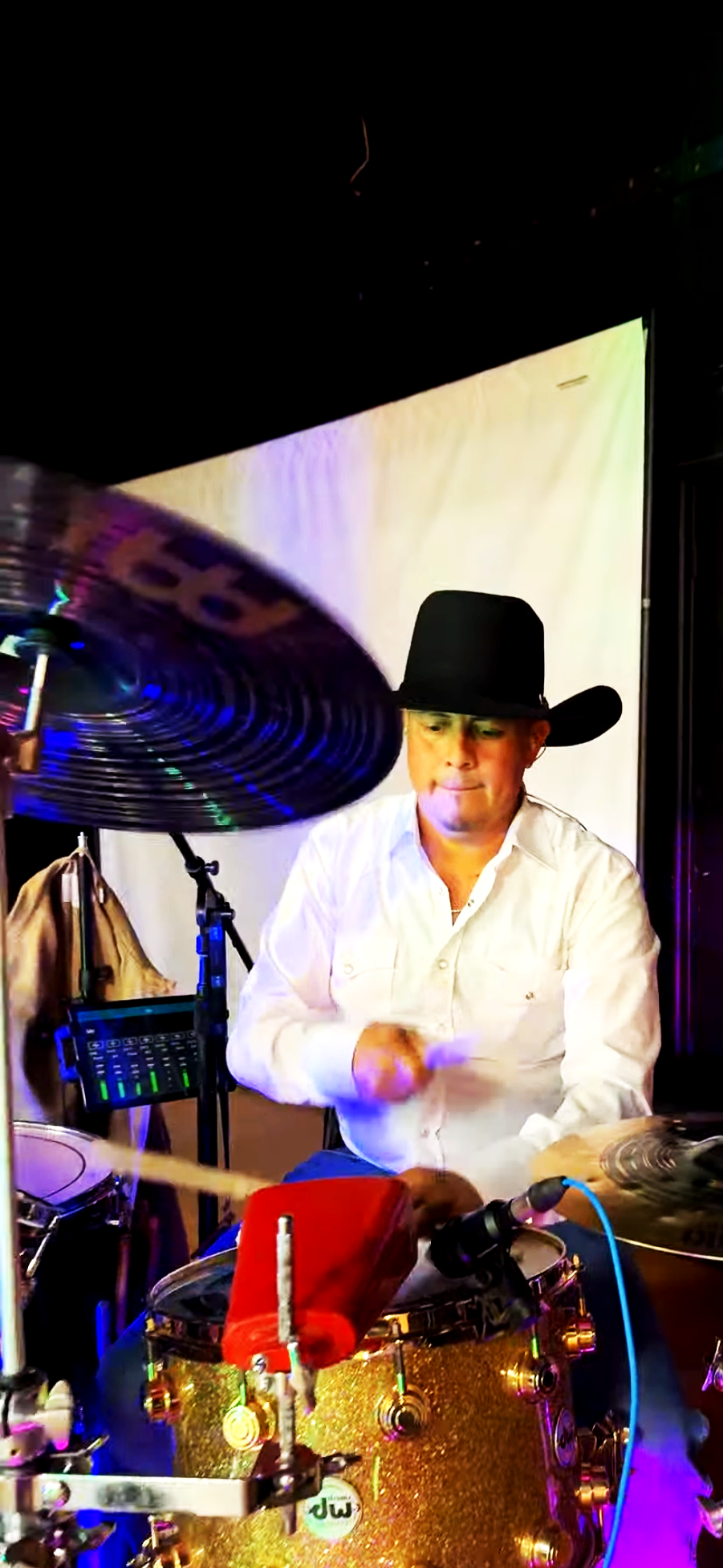 Hombre tocando la batería, lleva sombrero charro negro y camisa blanca, en un escenario con fondo de pantalla blanco y luces de colores.