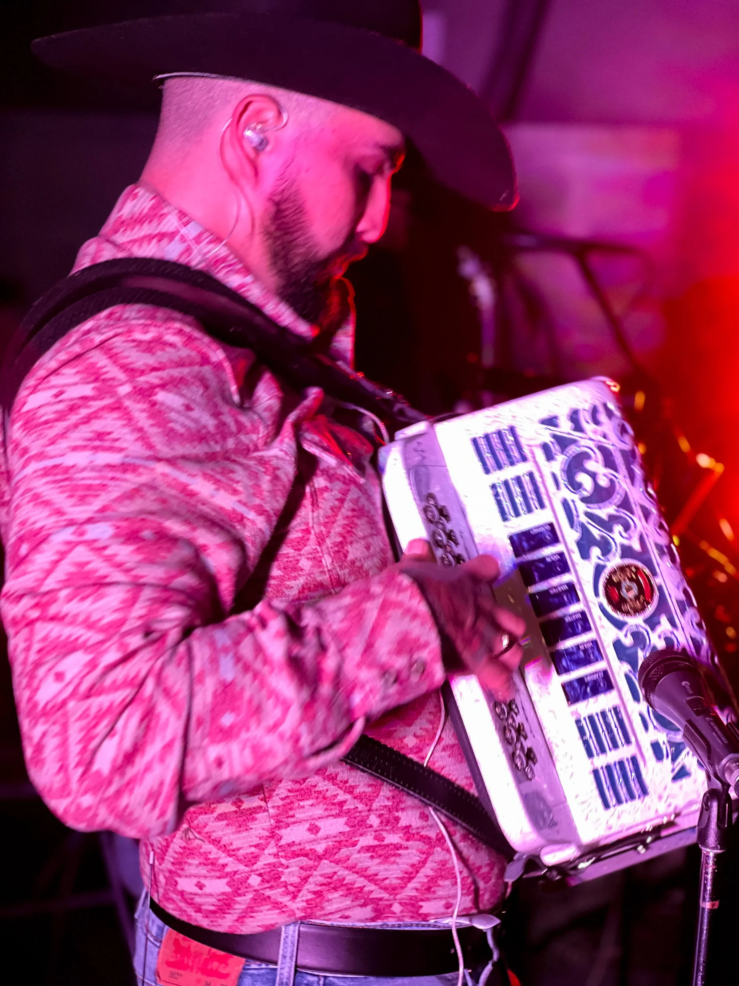 Persona con barba, ropa casual, usando auriculares y sosteniendo un mixer de DJ, en un ambiente con iluminación de color rosa y morado.