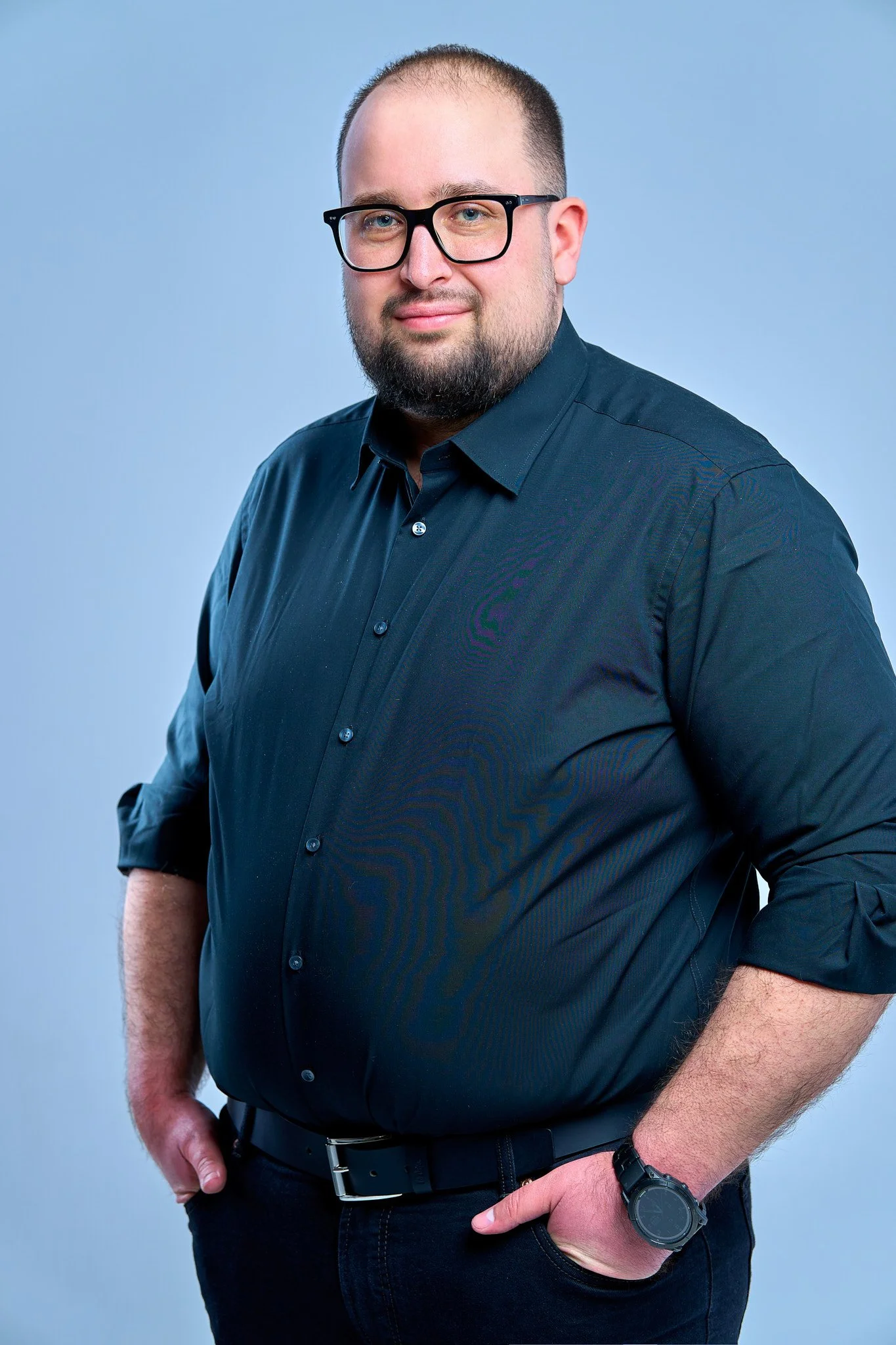 Portrait of a man wearing glasses and a dark button-up shirt, standing against a light blue background.