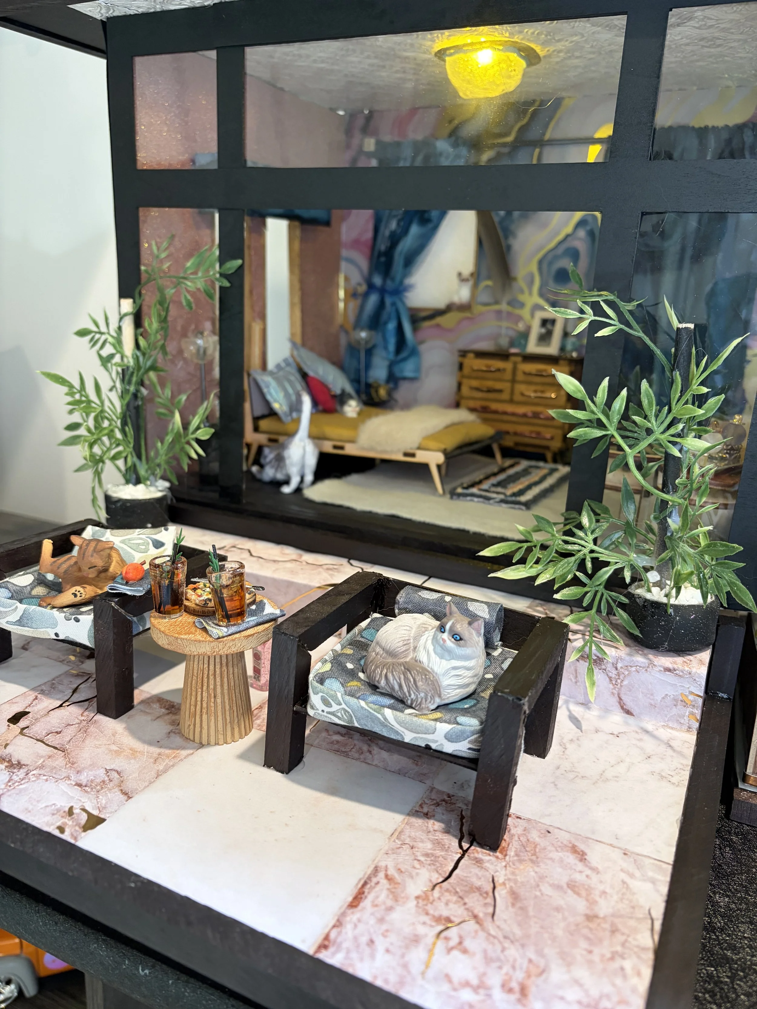 Miniature diorama of a cozy room viewed through a black window frame. The room features a bed with pillows, a blanket, a plush toy, and a bookshelf with a framed photo behind a blue curtain. There are two potted plants outside on a marble surface, an