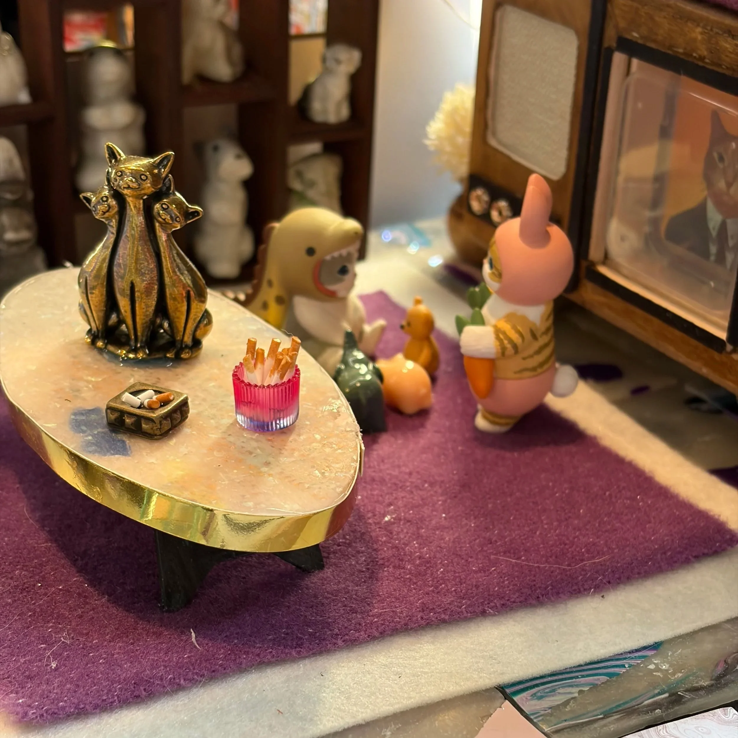 A small golden figurine of a cat with three smaller cats on its back, placed on a beige surface. In the background, there are various decorative items including a plush unicorn toy, shelves with figurines, and other ornaments.