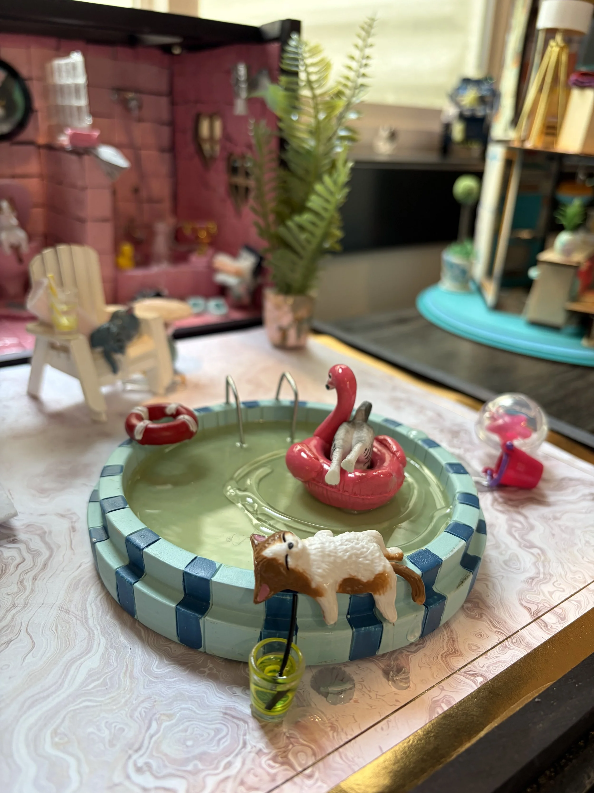 A small circular fountain with a tiger figurine on the edge, surrounded by decorative animals including a flamingo and a cat, placed on a table with a pinkish marble pattern in a cozy room with toys and plants in the background.