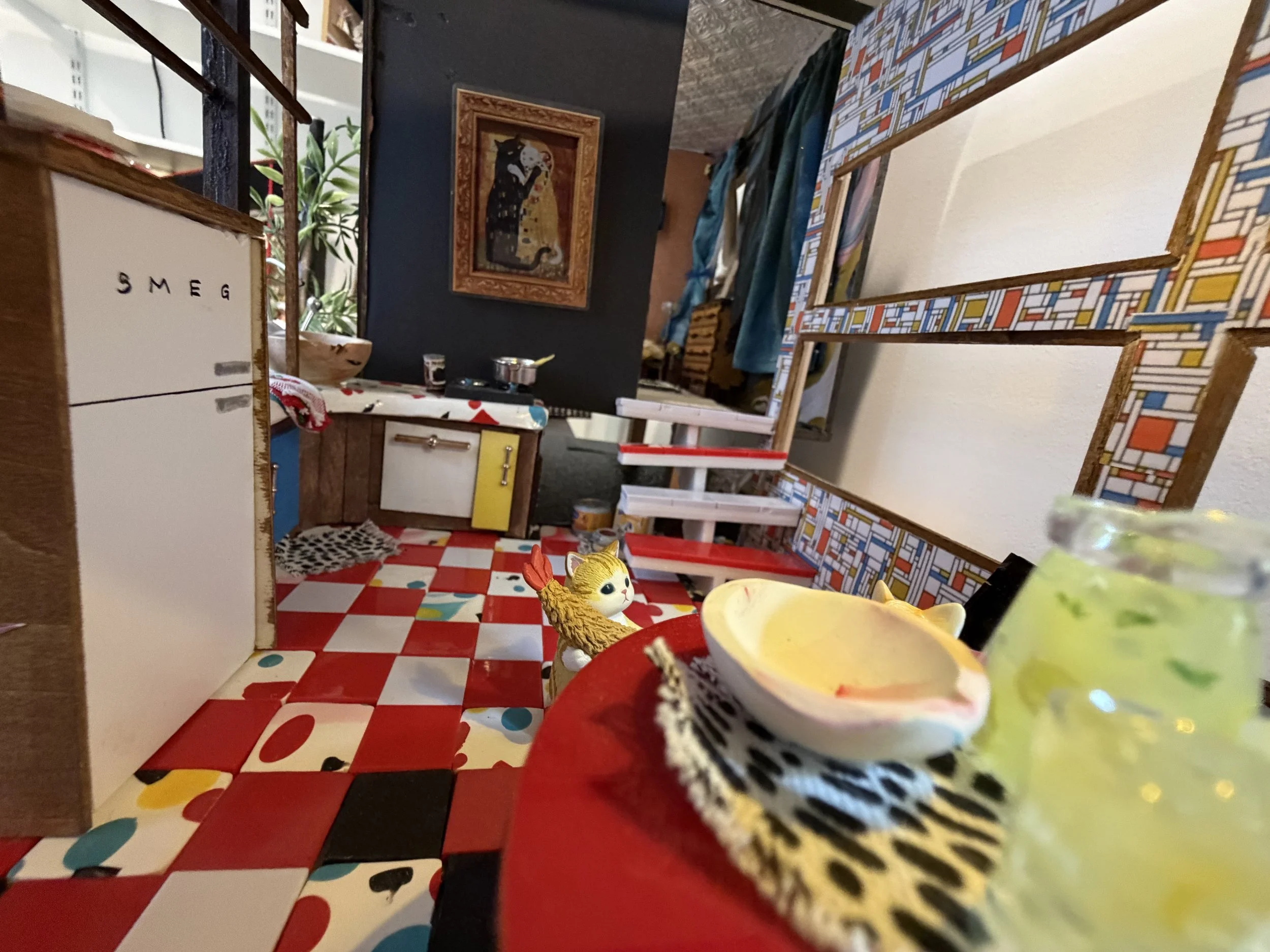 A colorful dining area with a red, white, and black checkered tablecloth, colorful chairs with geometric patterns, and various decorations including a framed artwork of a black cat, a small stuffed animal, and a potted plant. The background shows a k