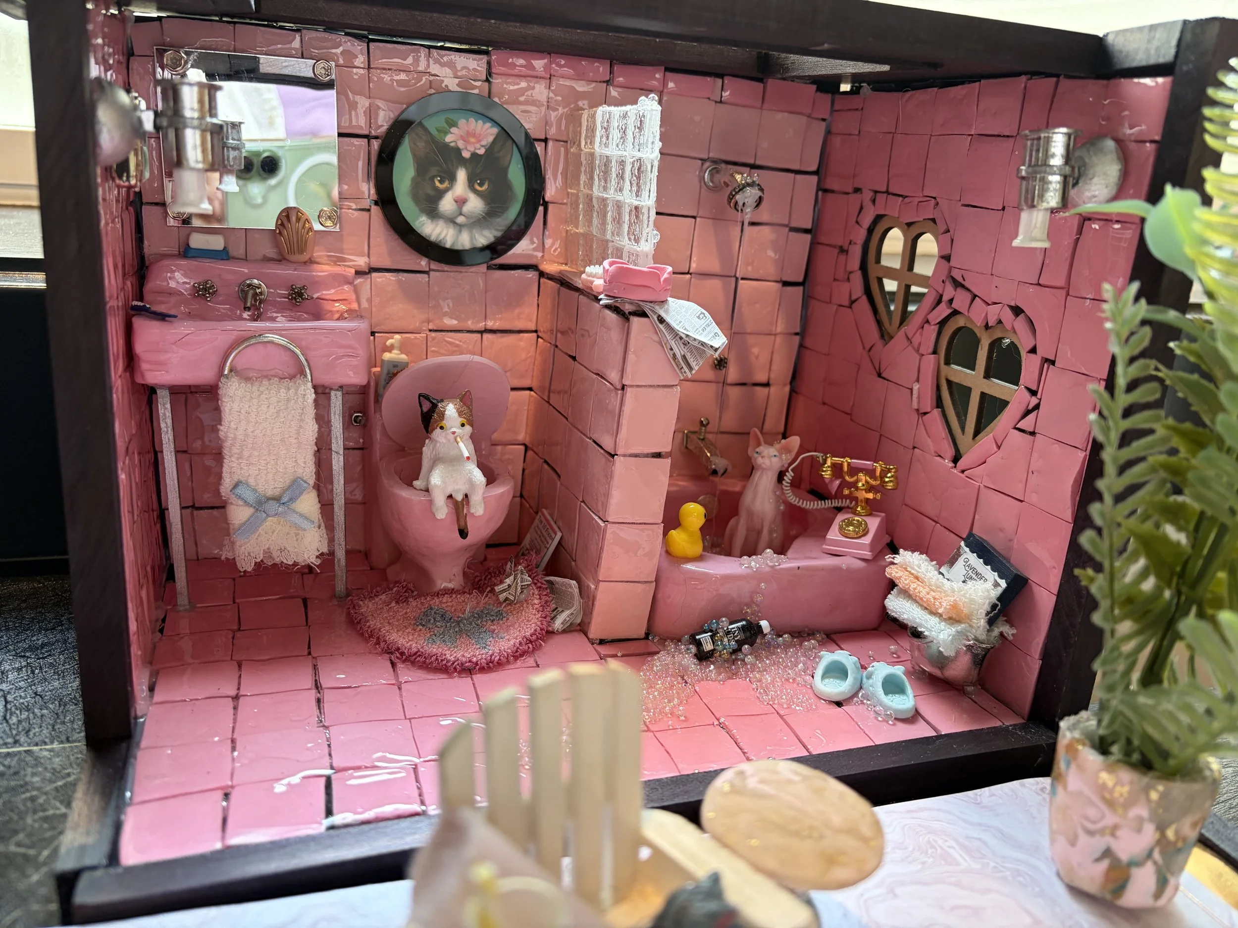 A miniature dollhouse bathroom decorated with pink bricks, featuring a cat-themed mirror, a small cat figurine on a pink toilet, and various tiny bathroom accessories and decorations.
