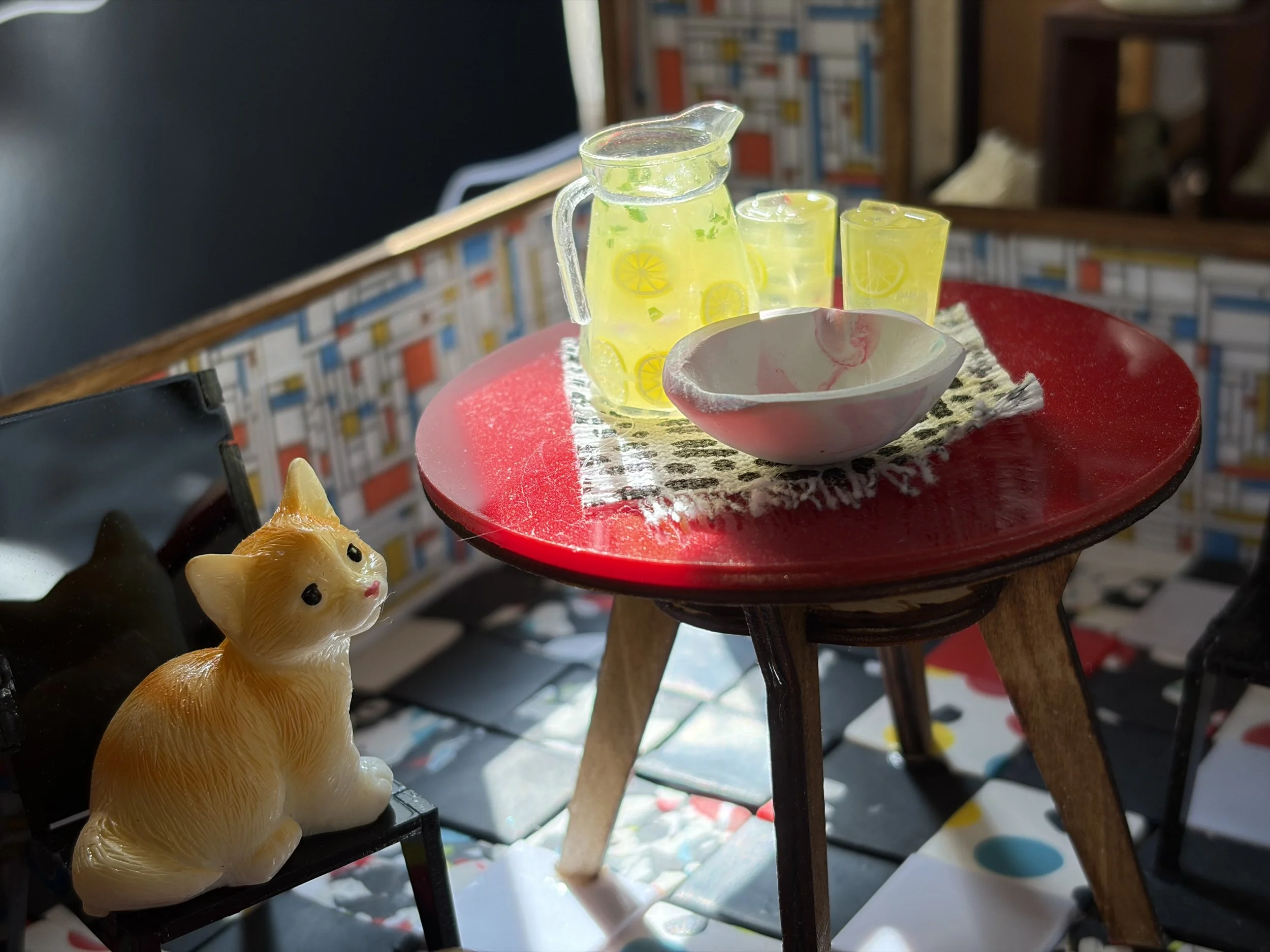 A glass pitcher and two glasses with lemon slices and green herbs, placed on a round red table with a red ceramic dish on top. A ceramic fox figurine is in the foreground. The background includes a bookshelf and a dark wall.