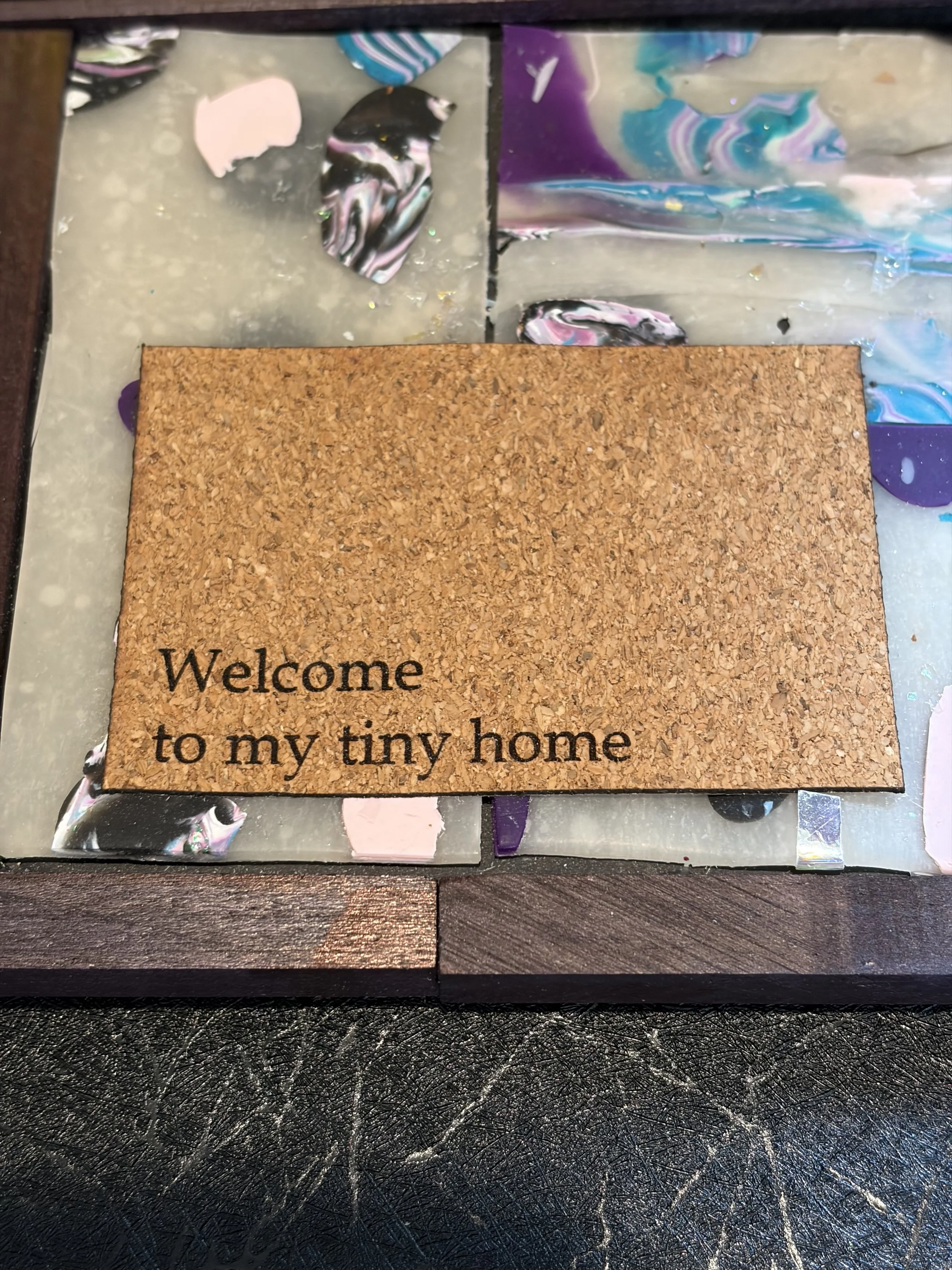 A small welcome mat with the text "Welcome to my tiny home" in black letters on a cork background, placed on a dark textured surface.