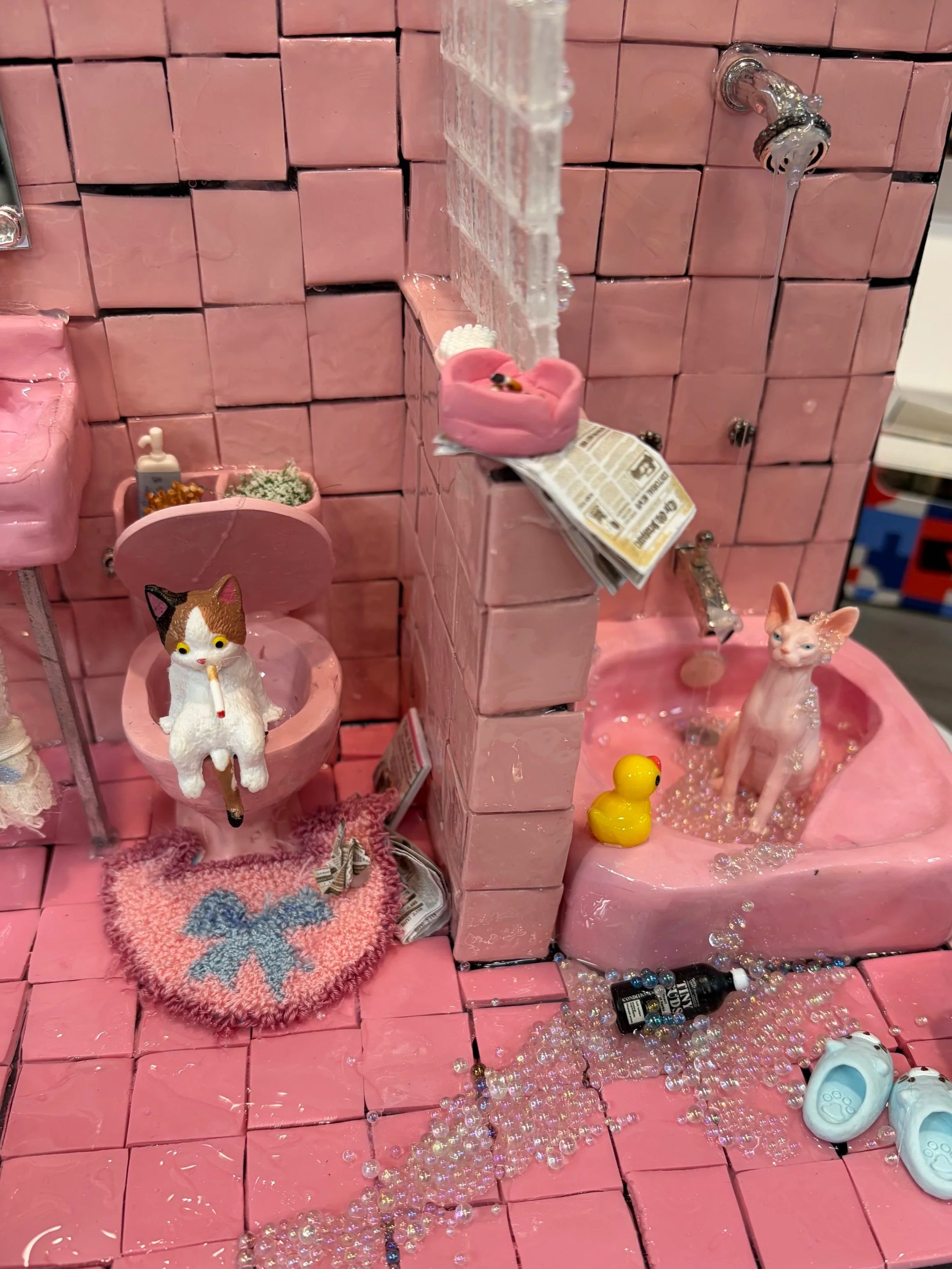 Decorative pink bathroom display featuring cat and dog figurines, pink tiled wall, small pink shelves with accessories, a rubber duck, and scattered beads and jewelry on a pink surface.