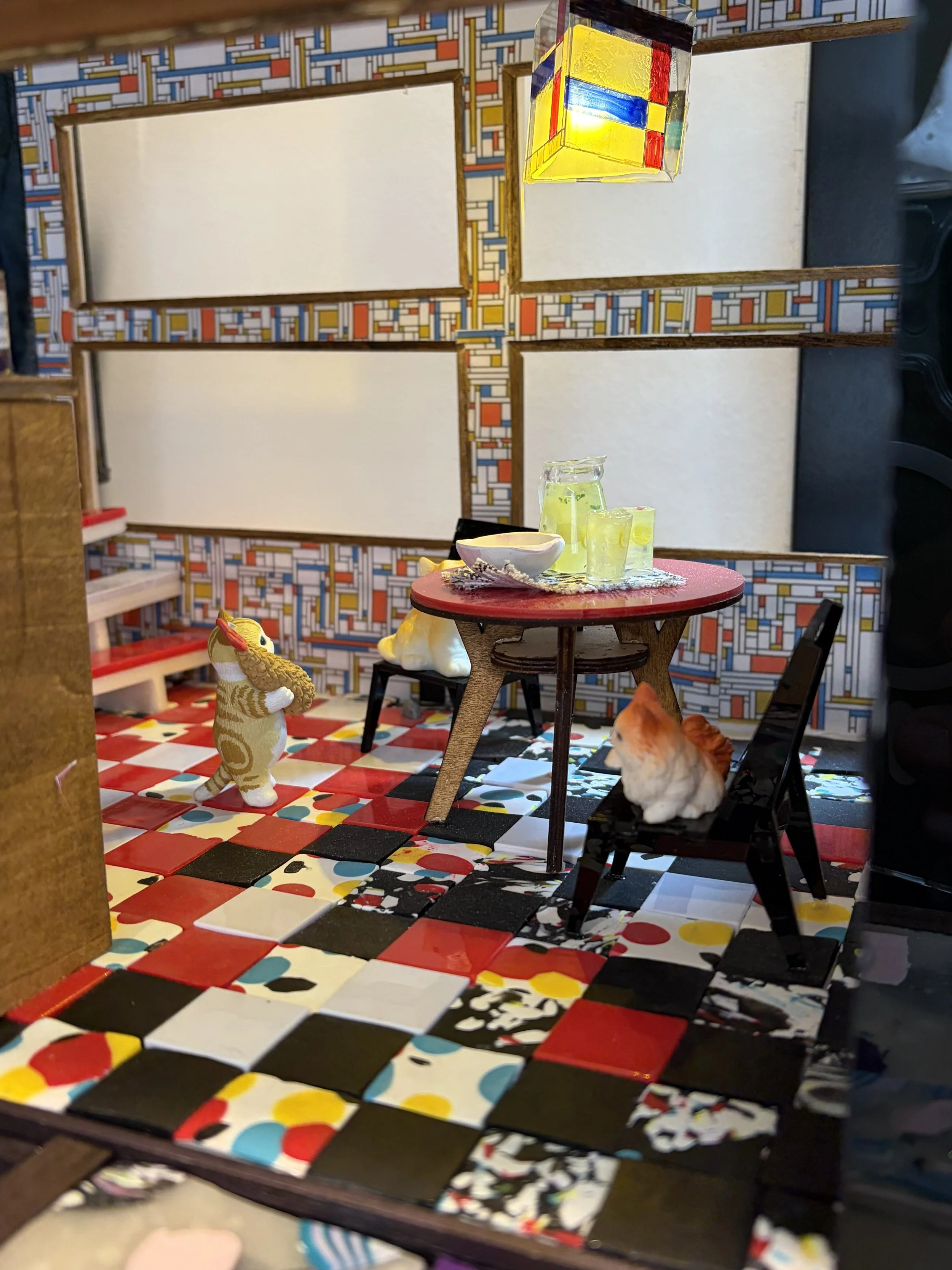 A miniature scene with a checkered red, white, black, and multicolored floor, a small round table with a red top and a cloth, a ceramic dog figure, a small white bowl, a glass pitcher filled with lemon water, and a ginger cat sitting near the table. 