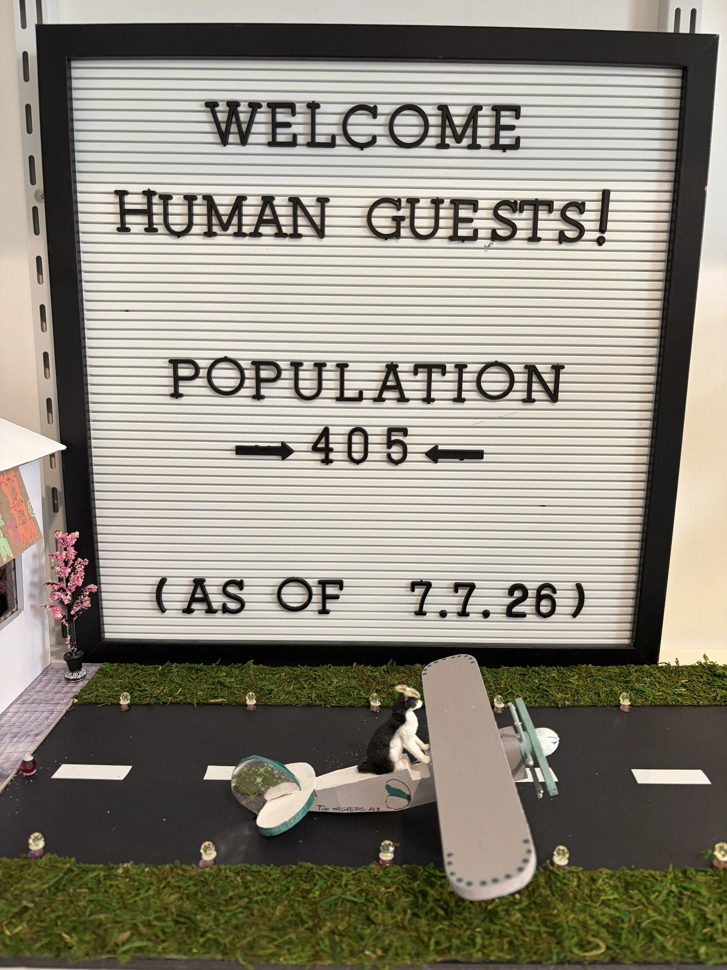 A welcome sign with black letters on a white background, displaying the text: "WELCOME HUMAN GUESTS! POPULATION 405 (AS OF 7.7.26)". In front of the sign, there are miniature figures including a black and white dog, a small decorative bush with pink 