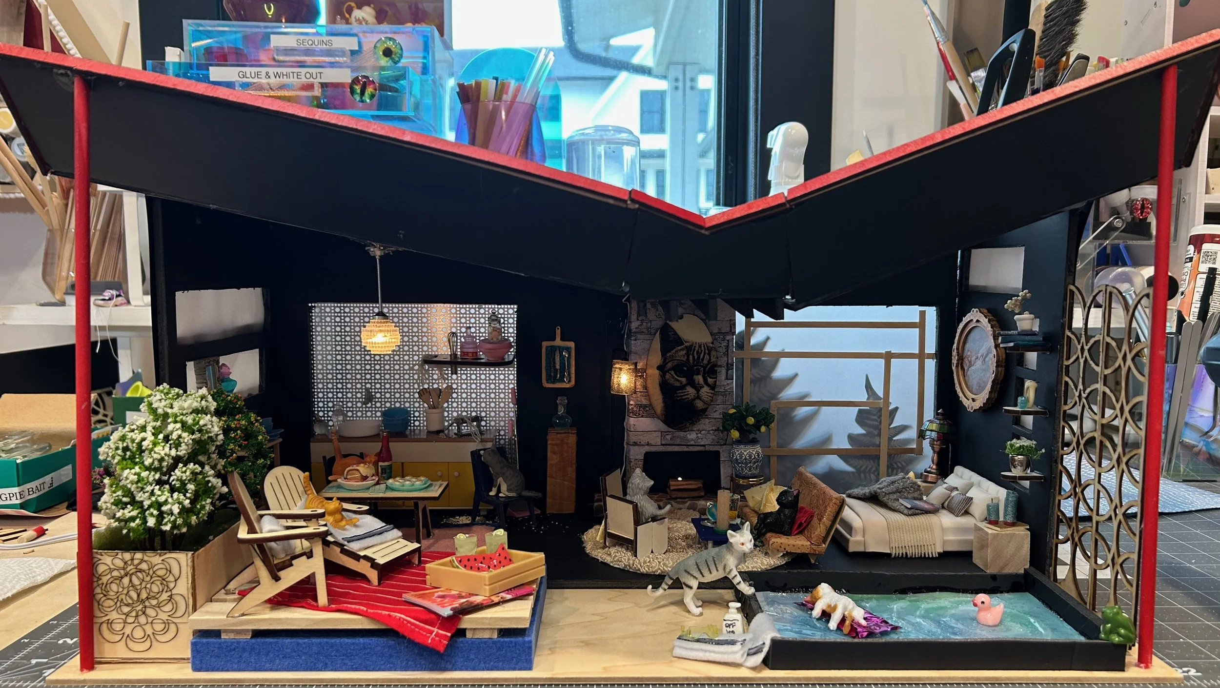Miniature dollhouse with detailed furniture and decorations, including a small dining table, chairs, plants, and miniature animals, set on a workspace with crafting supplies.
