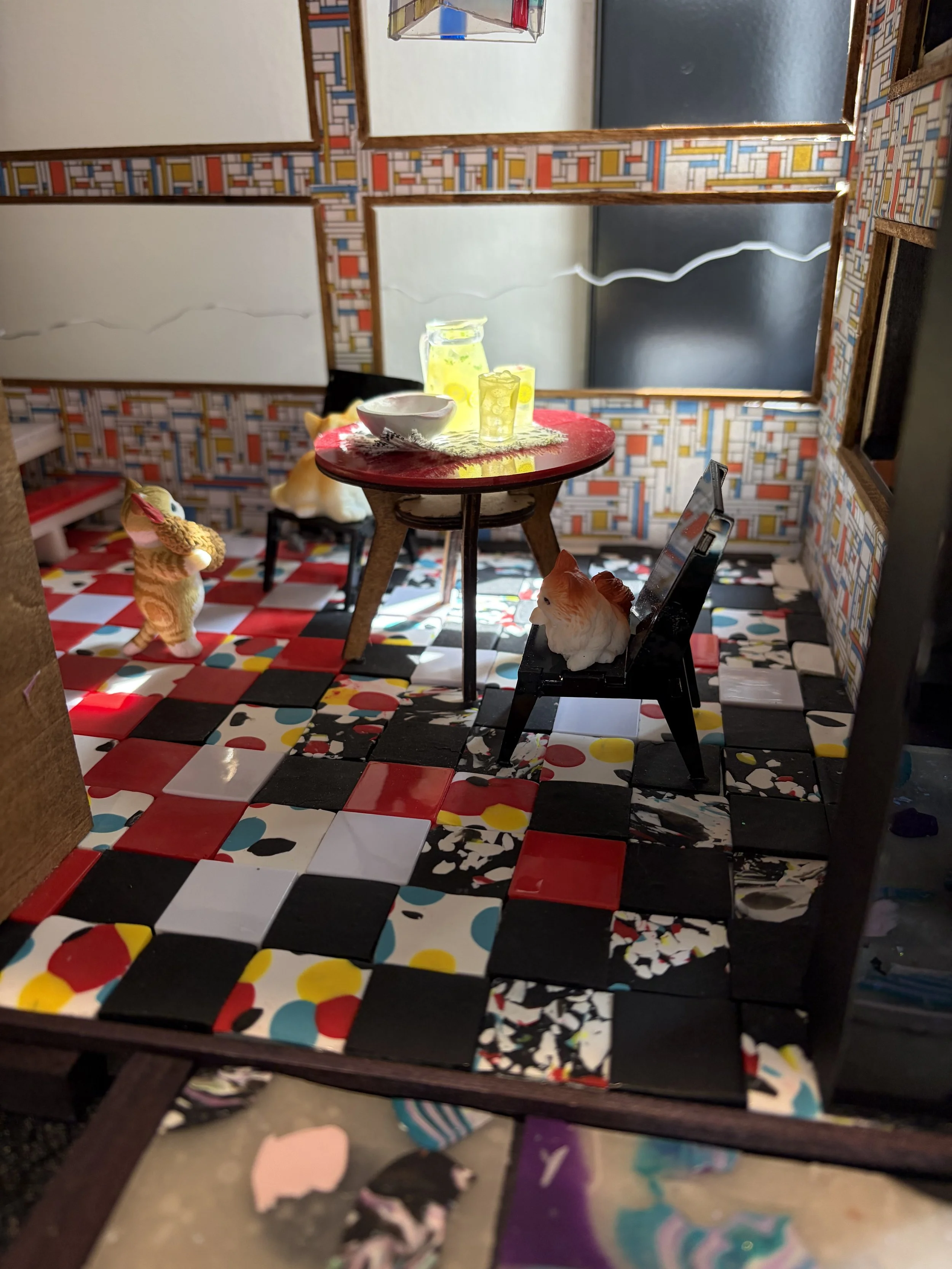 A miniature room with patterned wallpaper, a circus-themed floor, a small red table with a bowl and glasses, a mirror, and tiny animal figurines.