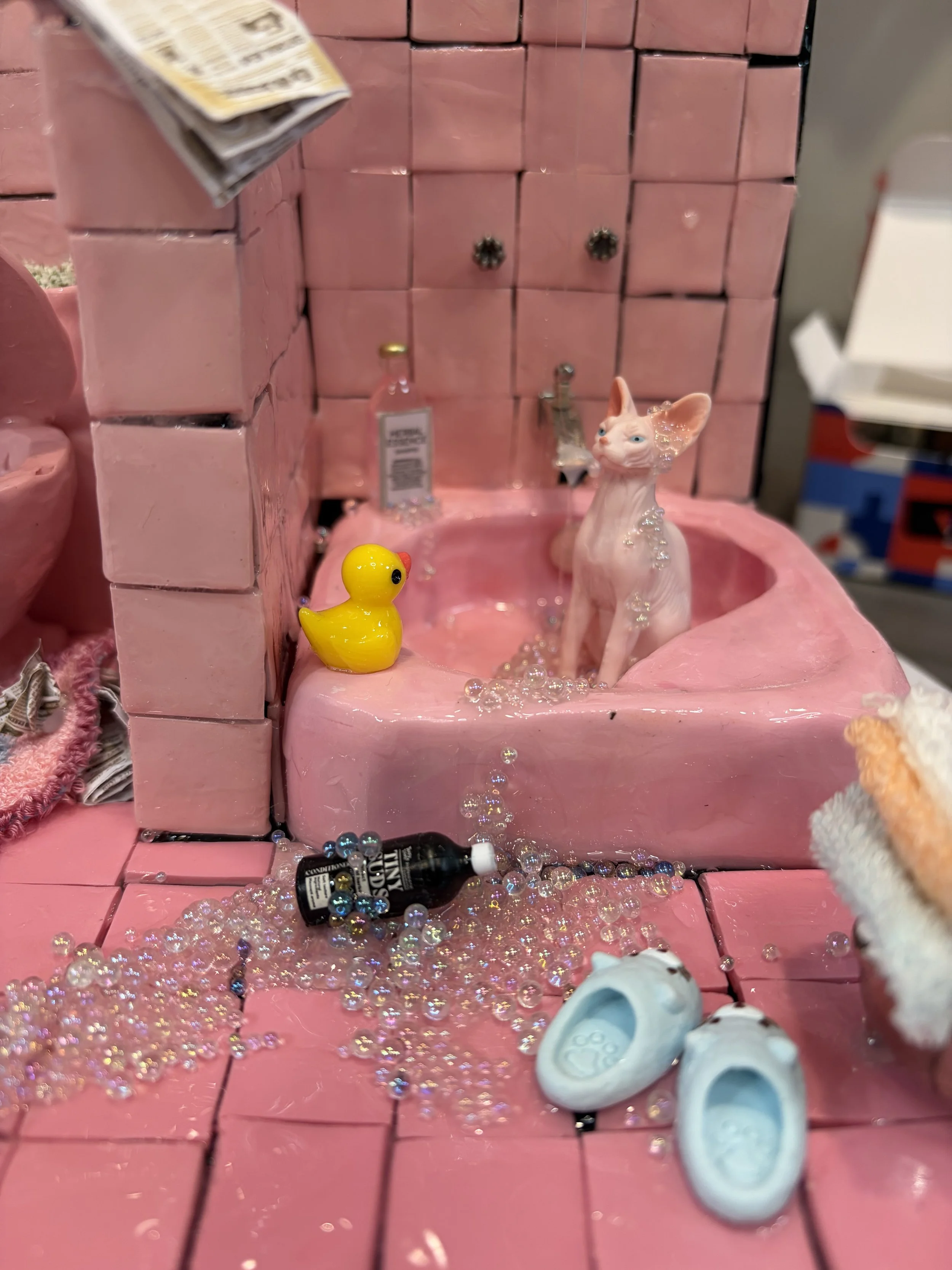 A miniature pink bathtub filled with water and small decorative items including a ceramic cat, a yellow rubber duck, a small bottle, and tiny beads and bubbles scattered around. White slippers are on the floor in front of the tub. The background has 