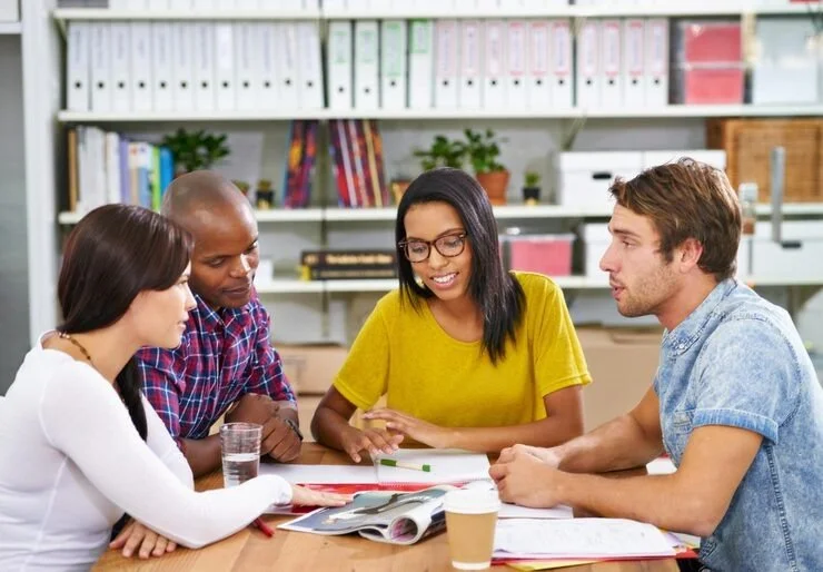 Four diverse young adults sit at a table engaged in discussion, with books, a notebook, a glass of water, a coffee cup, and a pen on the table, in a room with bookshelves.