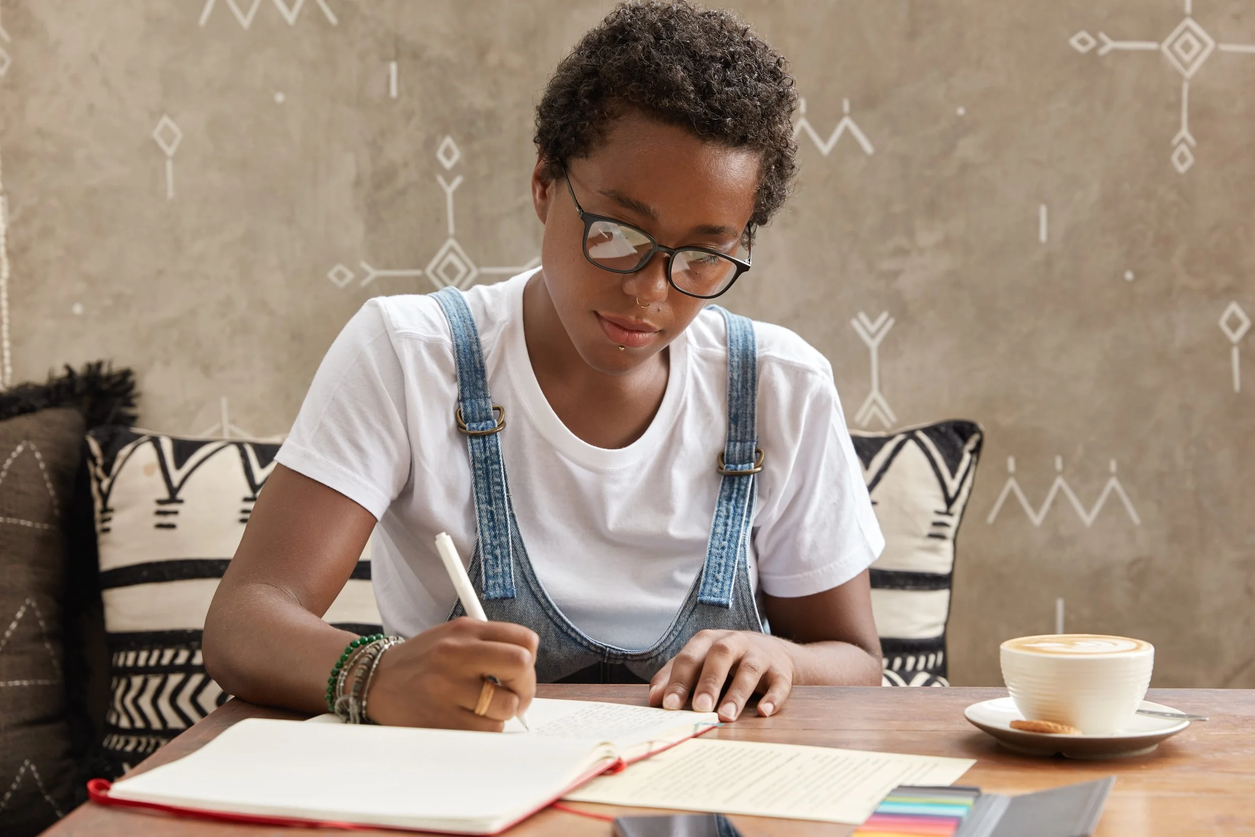 A young woman with short, curly hair and glasses, wearing a white t-shirt and denim overalls, sitting at a wooden table writing in a notebook. On the table, there is an open book and a cup of coffee or tea. The background features a patterned wall and a decorative pillow on a sofa.