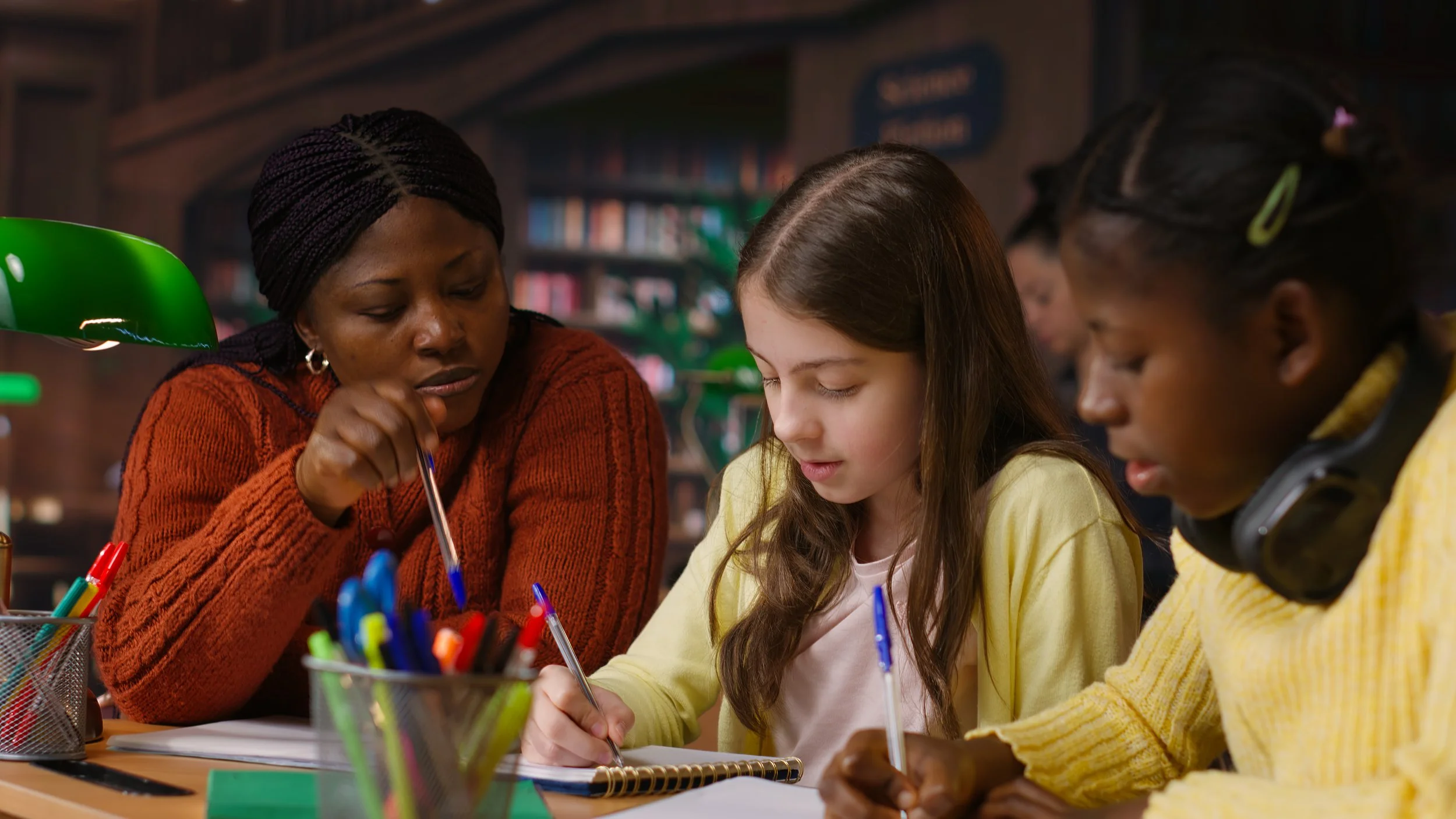 An adult and two children sit at a table in a library or study area, focusing on writing or drawing with pens. There are colorful pens in containers on the table, and bookshelves are visible in the background.