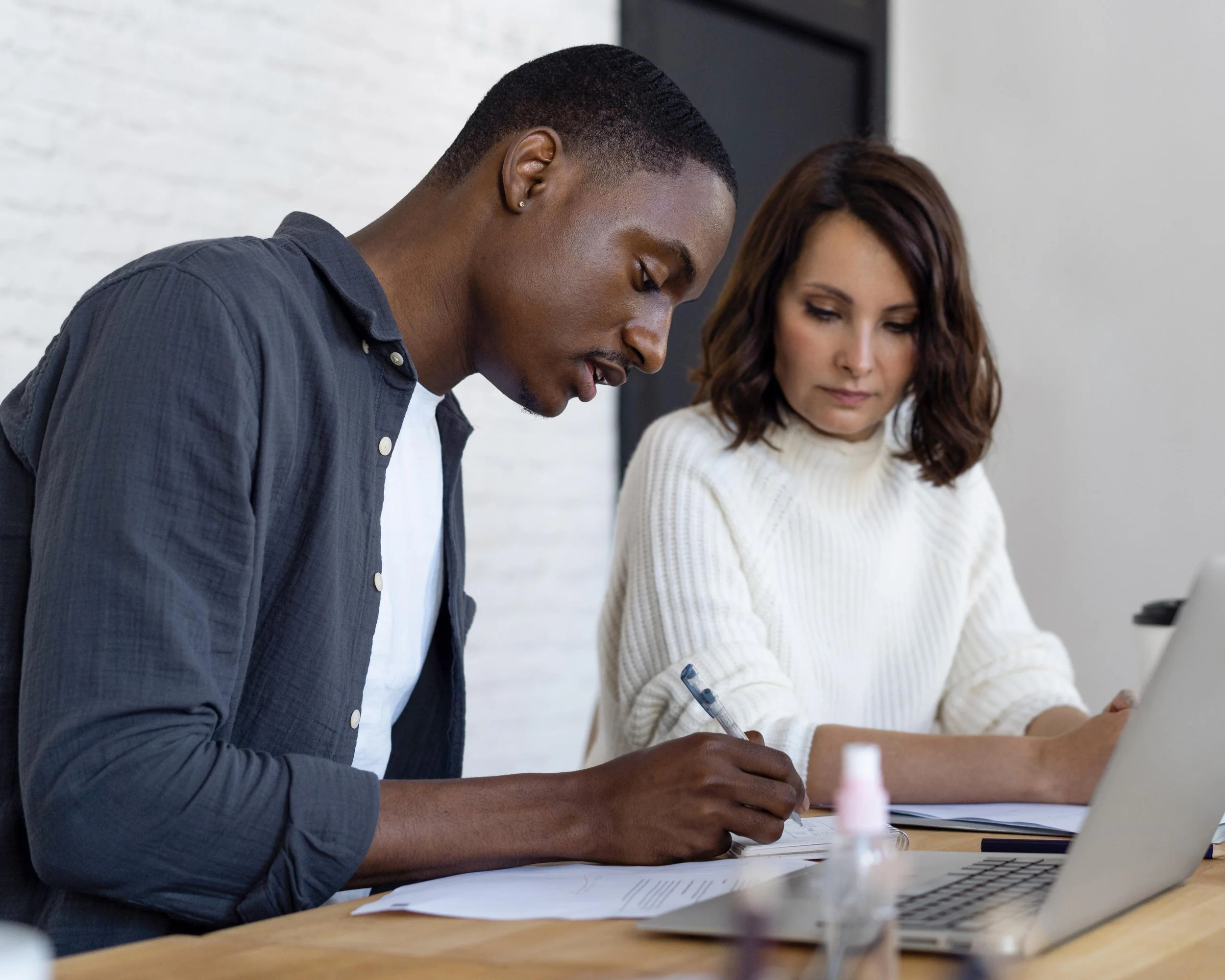 A man with dark skin and short black hair is writing in a notebook, sitting across from a woman with light skin and wavy brown hair, who is looking down and possibly also writing. They are in a room with a white brick wall.