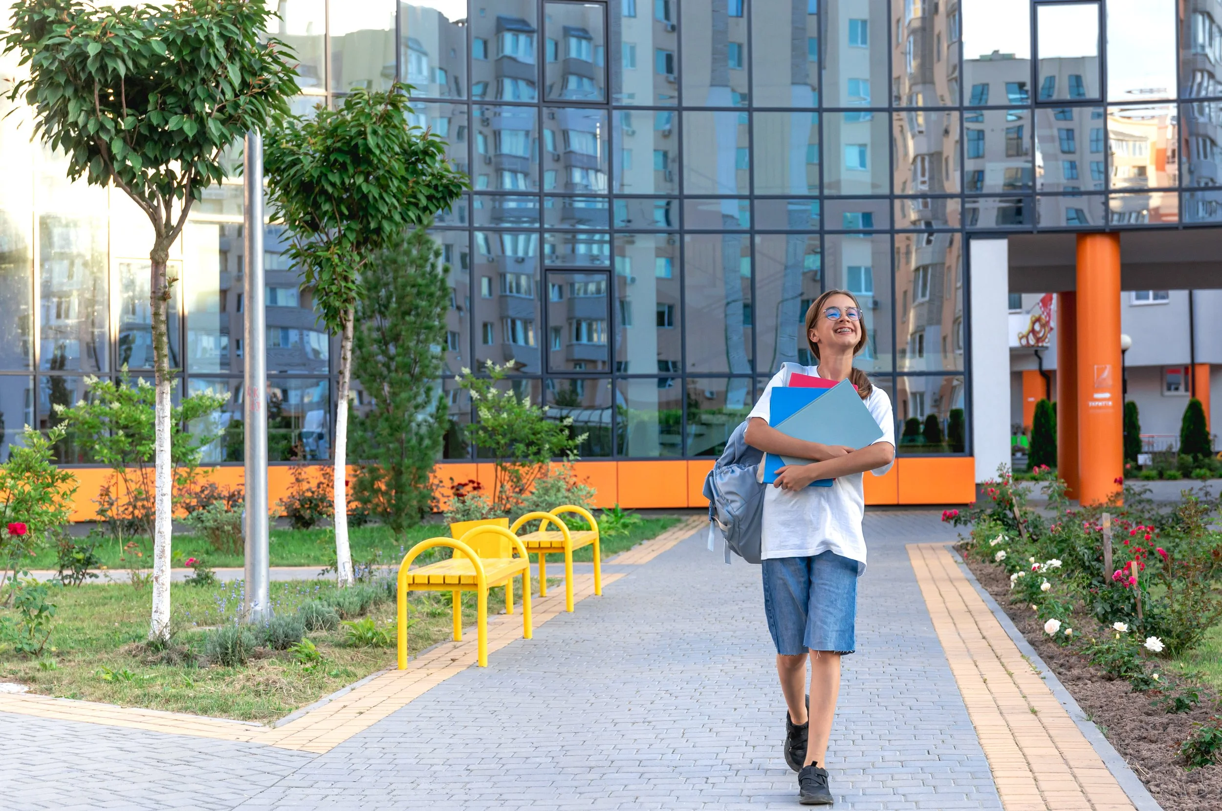 A young girl wearing glasses, a white T-shirt, and jeans walking on a paved pathway outside a modern glass building surrounded by trees and colorful flowers, holding folders and a backpack.