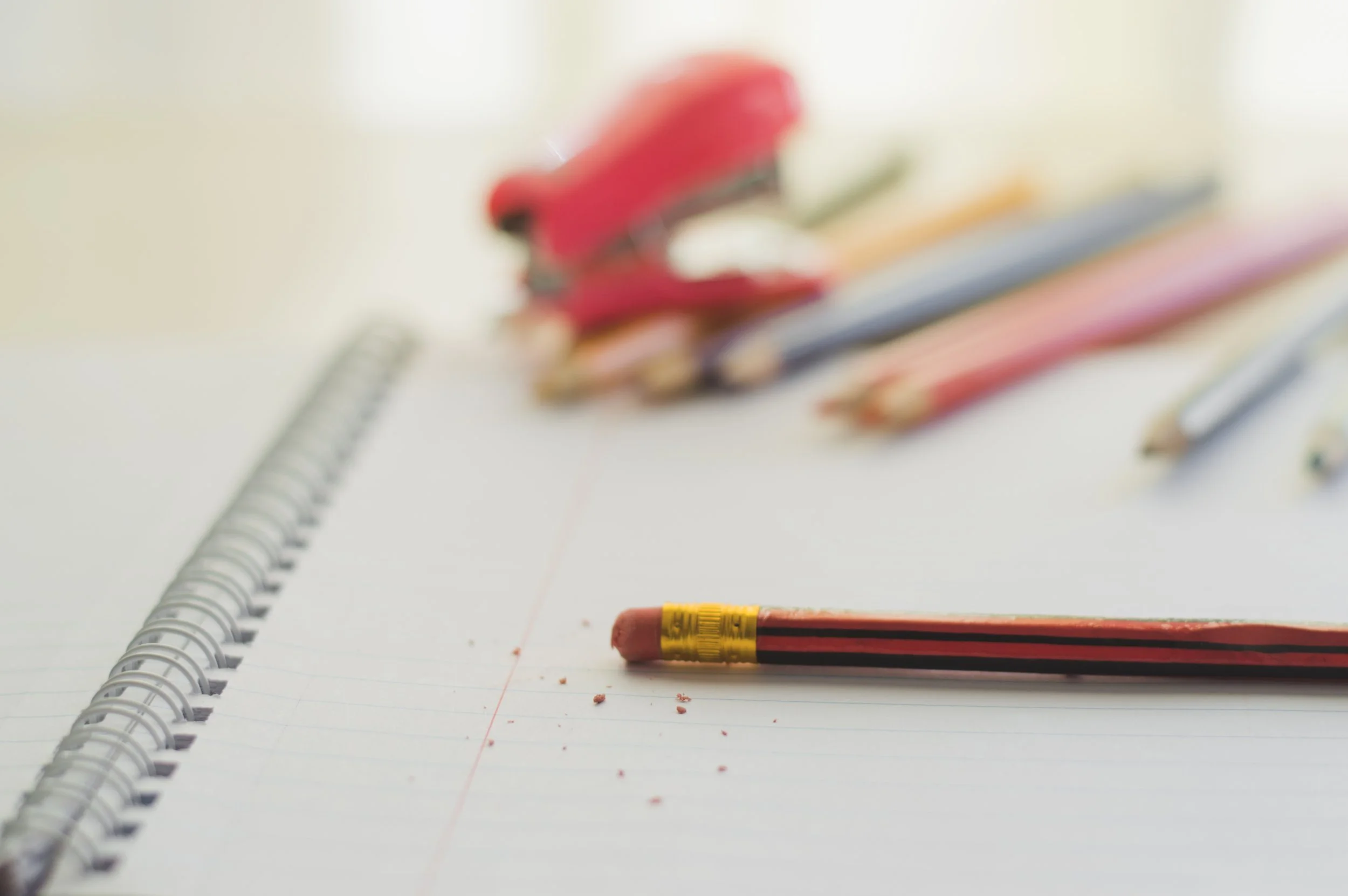 Open notebook with red and yellow pencil and a pen on a white background, slightly out of focus.
