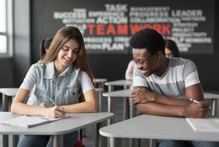 Two students sitting at desks in a classroom, smiling and working on assignments. In the background, a poster with words like 'teamwork' and 'success' is visible.