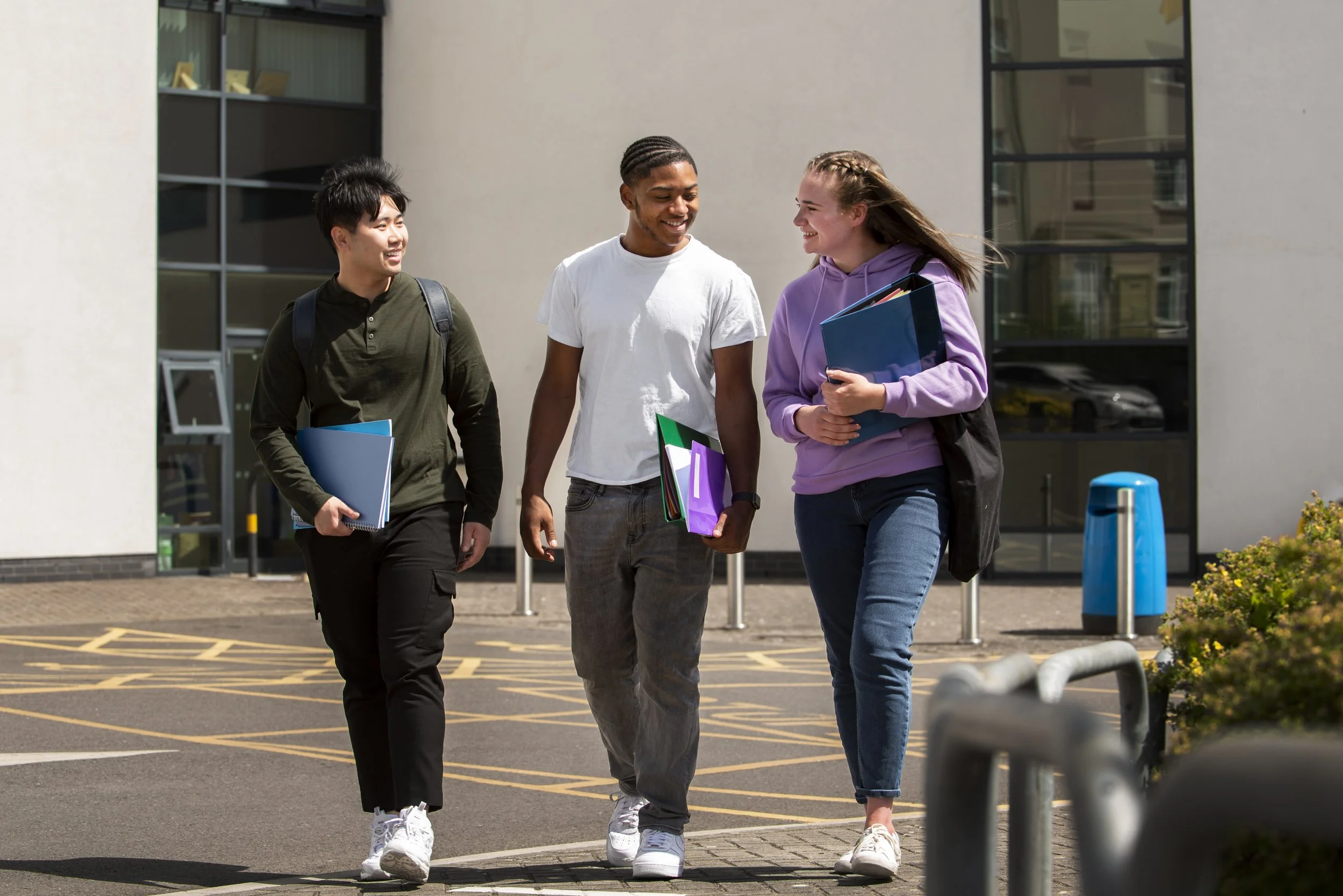 Three college students walking outside, carrying books and backpacks, smiling and chatting.
