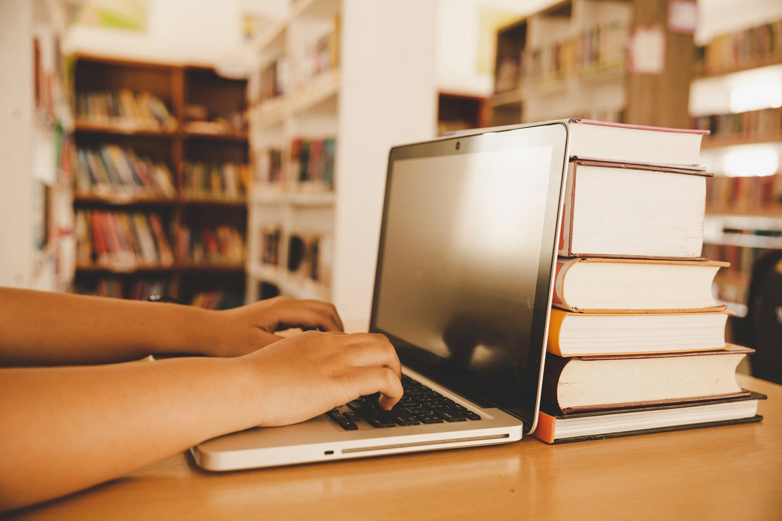 A person using a laptop at a desk with a stack of books nearby in a library.