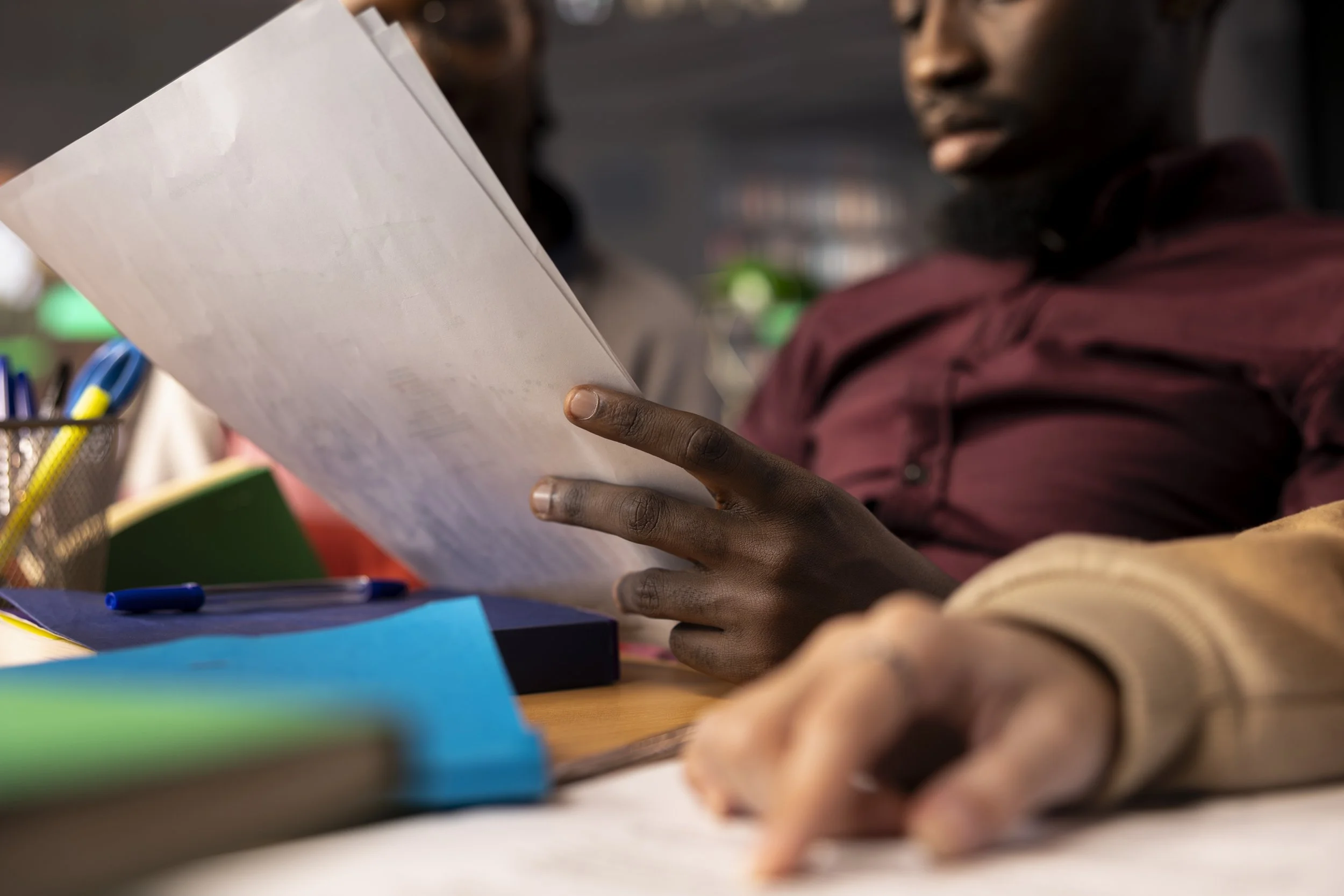 A person holding a white piece of paper or notebook, with colorful folders and writing utensils on a desk, in a setting that appears to be a classroom or office.