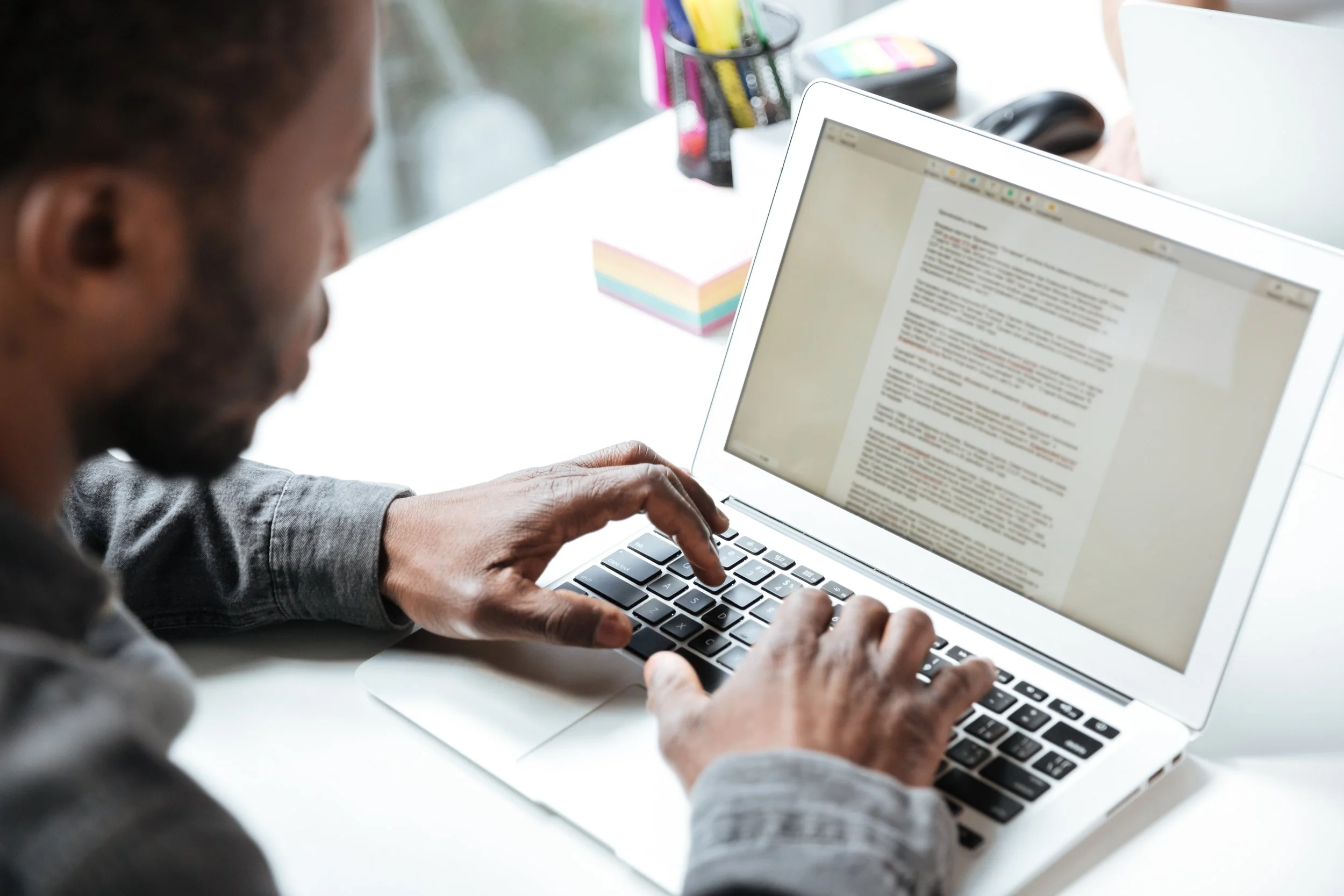 A person with dark curly hair and a beard typing on a MacBook laptop at a white desk. The screen displays a document with highlighted text. In the background, there are colorful sticky notes, a black container with various pens and markers, a black computer mouse, and a closed black case.