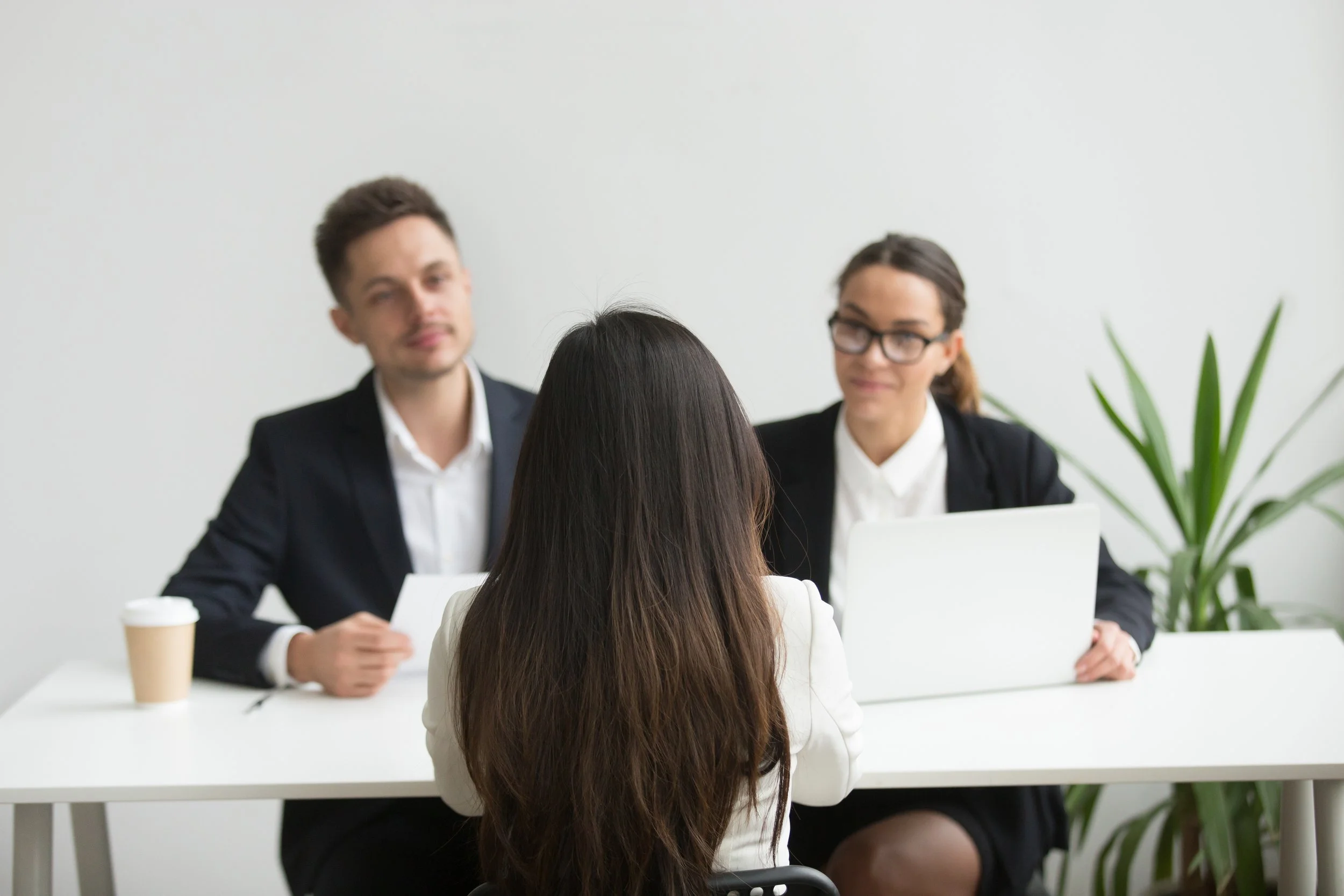 Four people in a business meeting, two women and two men, sitting around a white table in an office with a white wall and green plant visible. The woman in the foreground has long dark hair and is facing away from the camera. The man and woman in the background are looking towards her.