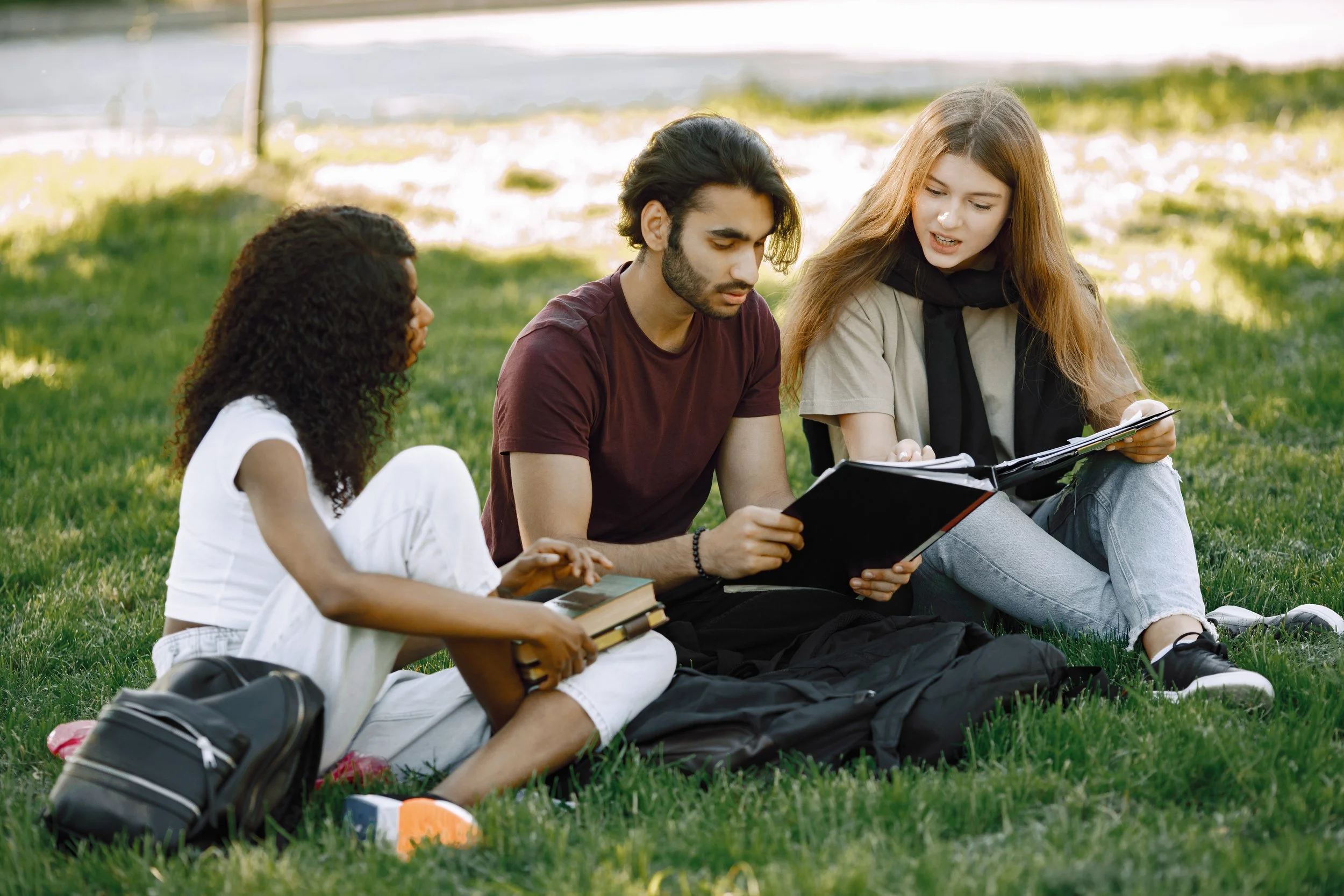 Group of four young people sitting on grass outdoors, looking at a book and a phone, with backpacks nearby.