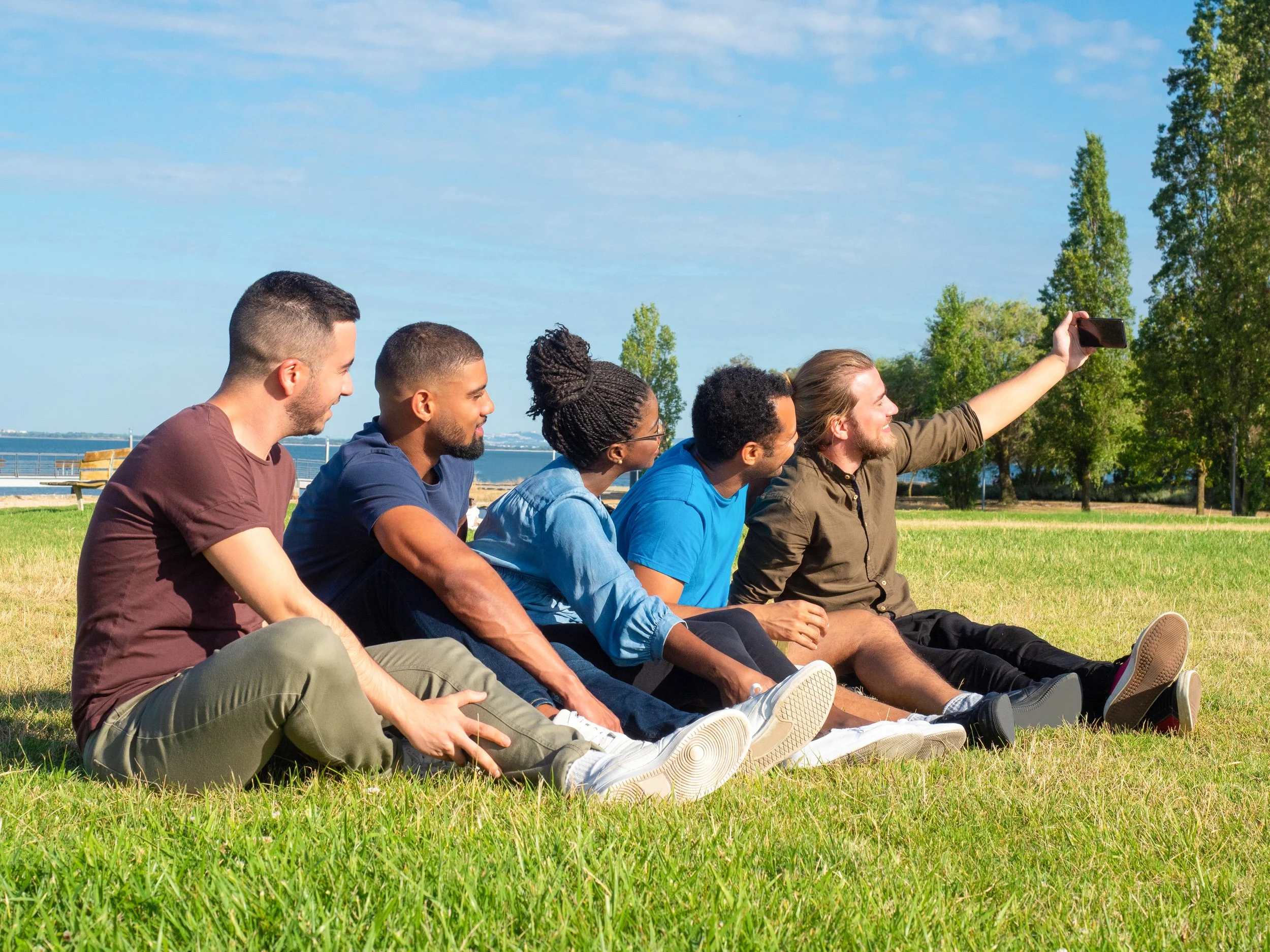 Group of six friends sitting on grass in a park taking a selfie with a smartphone, with trees and a body of water in the background.