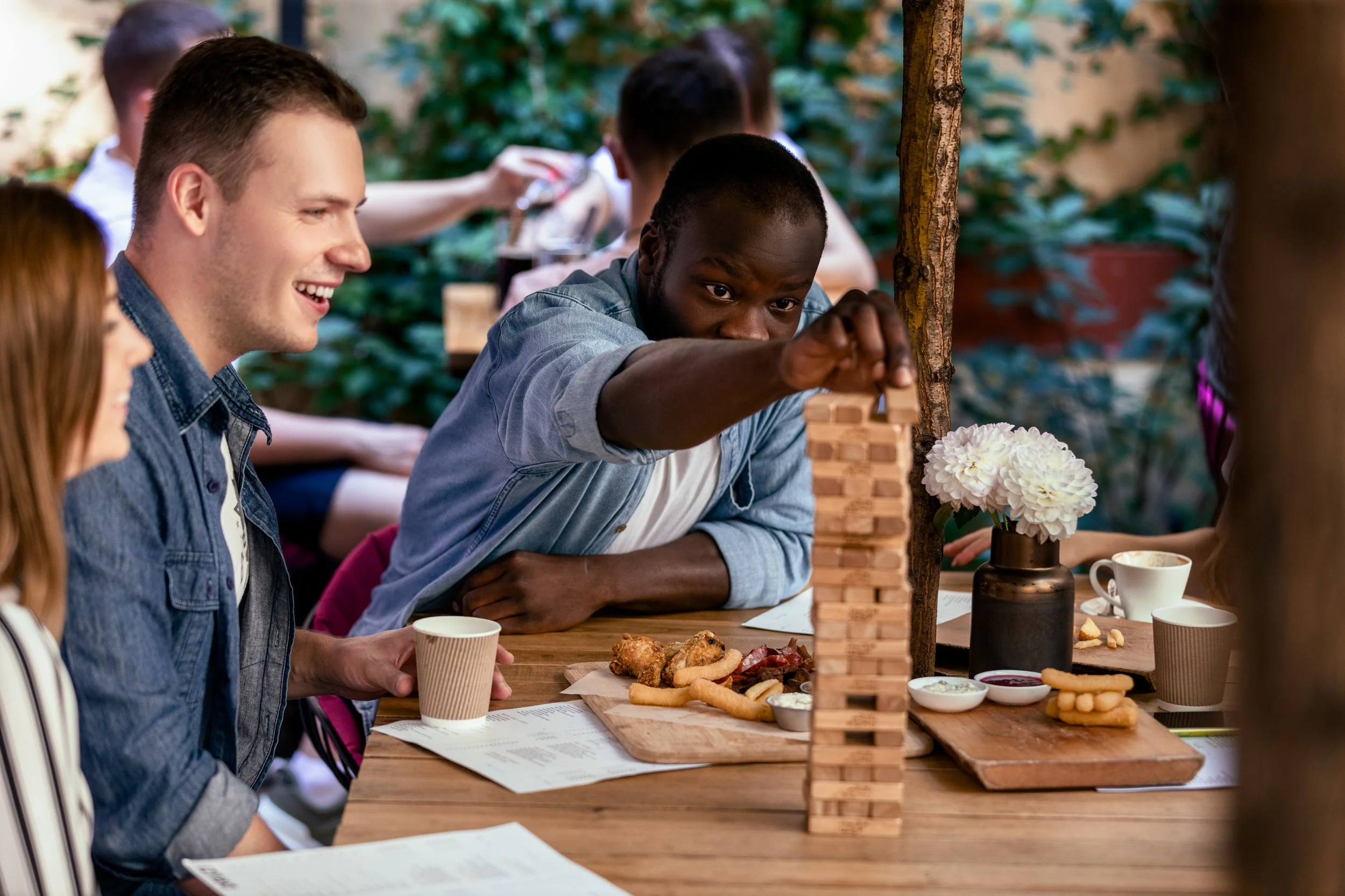 Group of people playing a giant Jenga game at a wooden table during a meal outdoors. There are various snacks and drinks on the table, including fried chicken, cookies, and cups. The setting is surrounded by greenery.