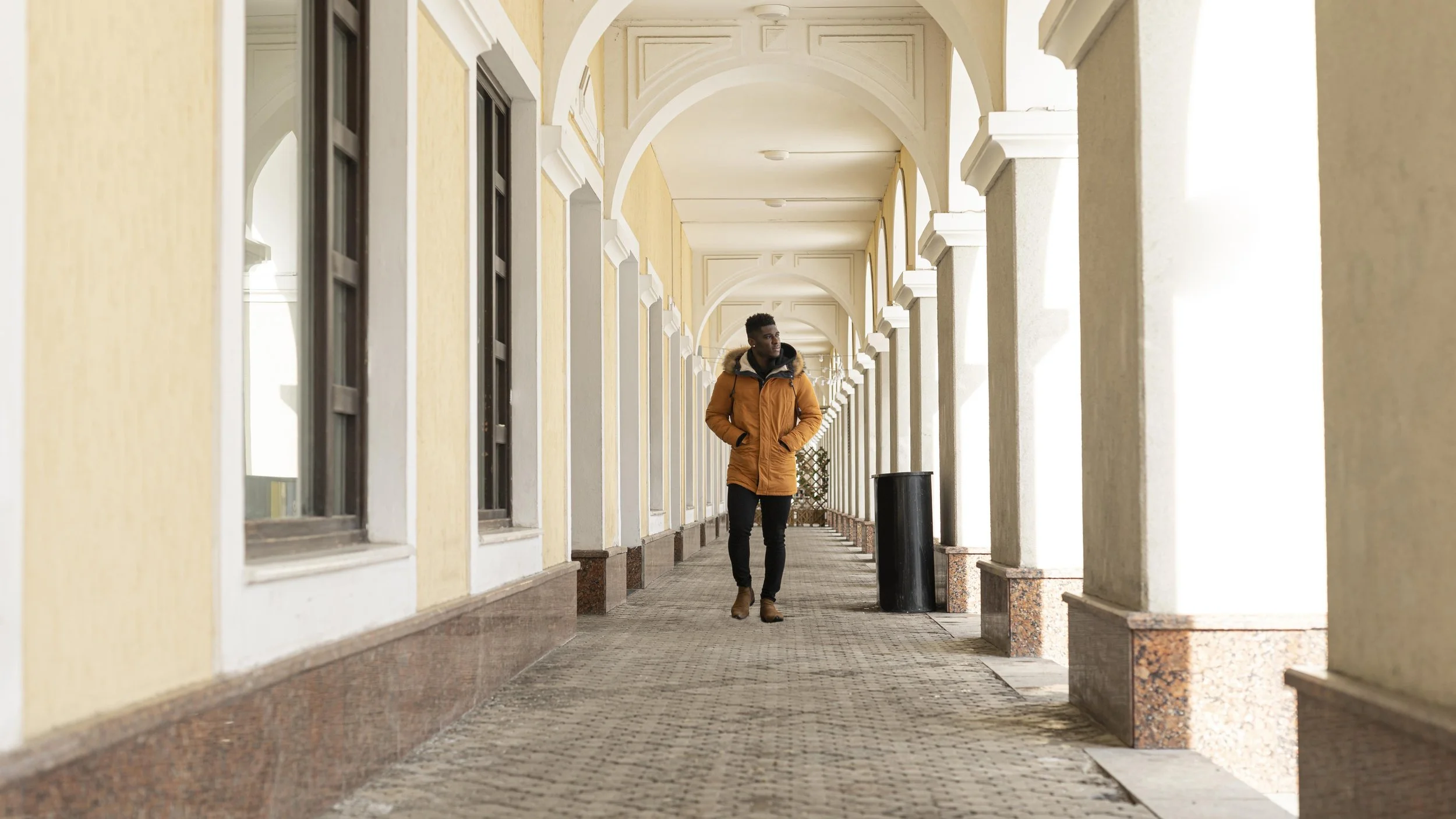 A man walking through a covered outdoor walkway with yellow walls, arched ceilings, and large windows, wearing a brown jacket and black pants.