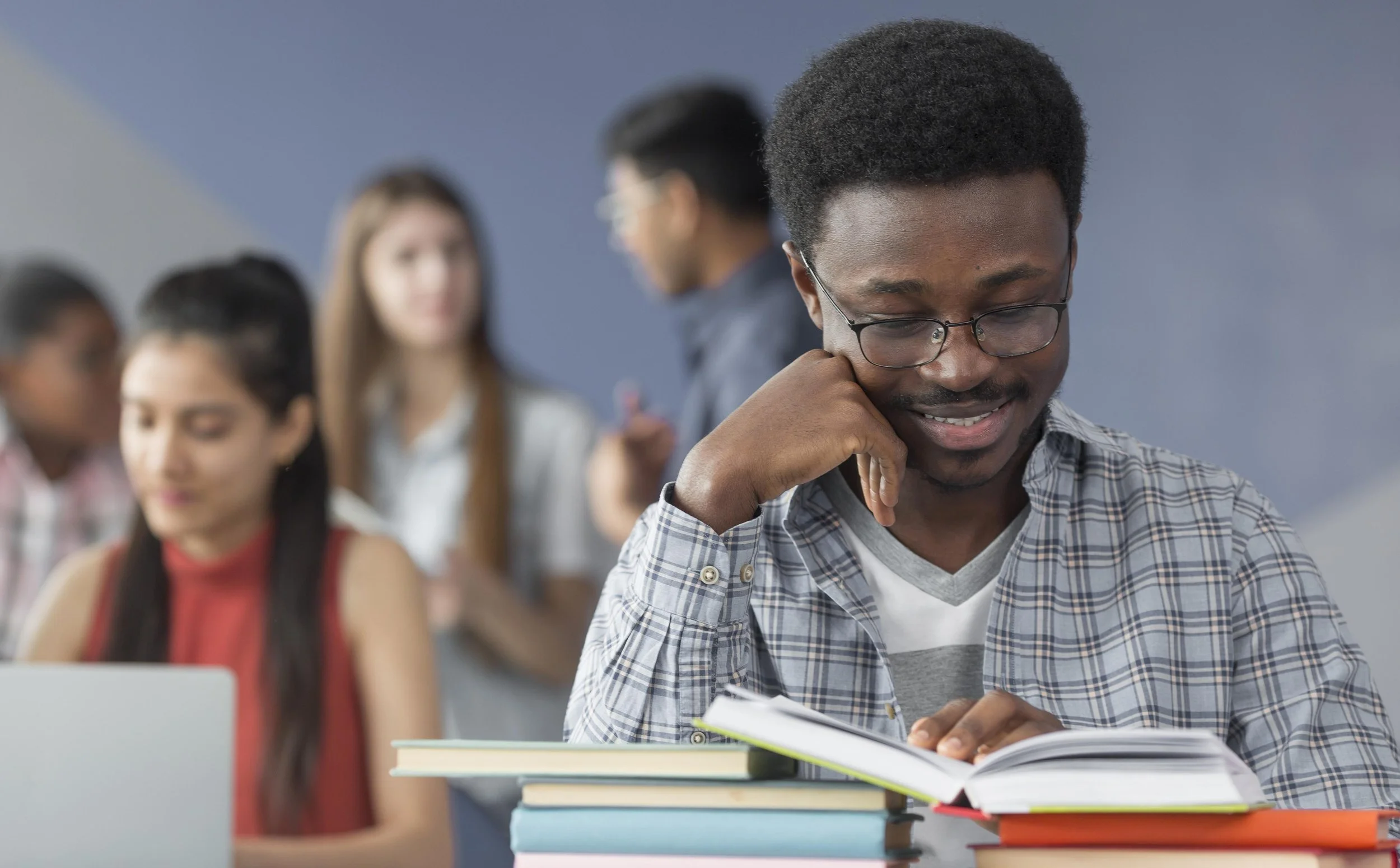 A man wearing glasses and a plaid shirt, studying a book, with a stack of books in front of him, in a classroom with other students in the background.