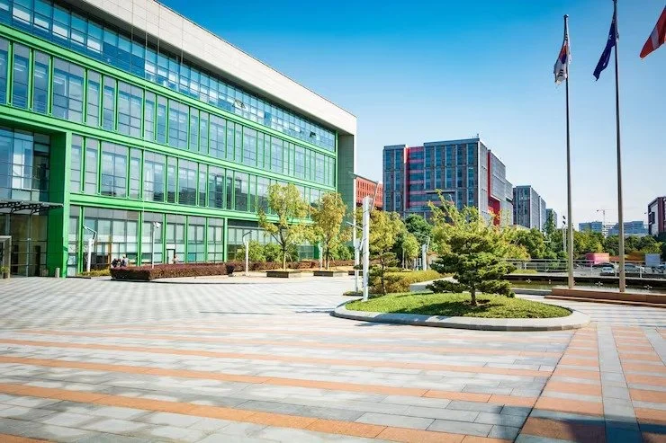 Modern office building with green glass facade, flagpoles with flags, trees, and paved plaza area under a clear blue sky.
