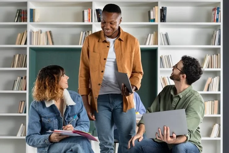 Three young adults in a library or study room, two sitting and one standing, engaged in conversation. The woman on the left holds a notebook and pen, the man on the right holds a laptop, and the man standing has a tablet in his hand, all smiling and interacting.