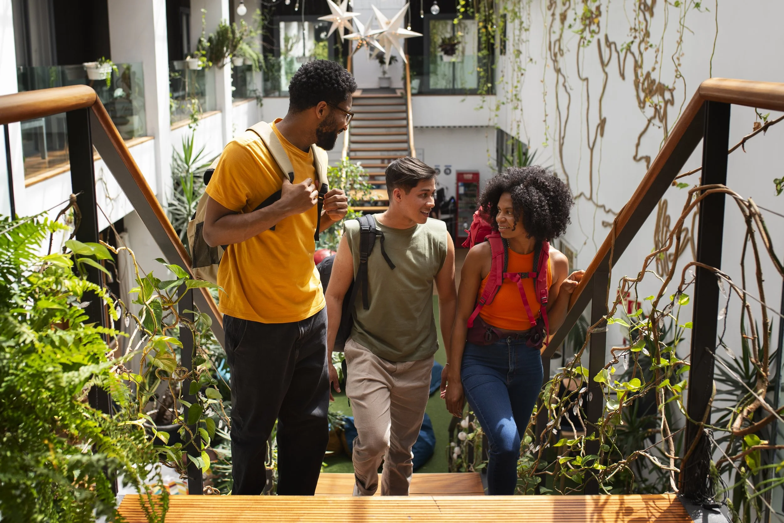 Three young adults with backpacks walking up a staircase in a green indoor garden or greenhouse, surrounded by various plants and decorative hanging stars.