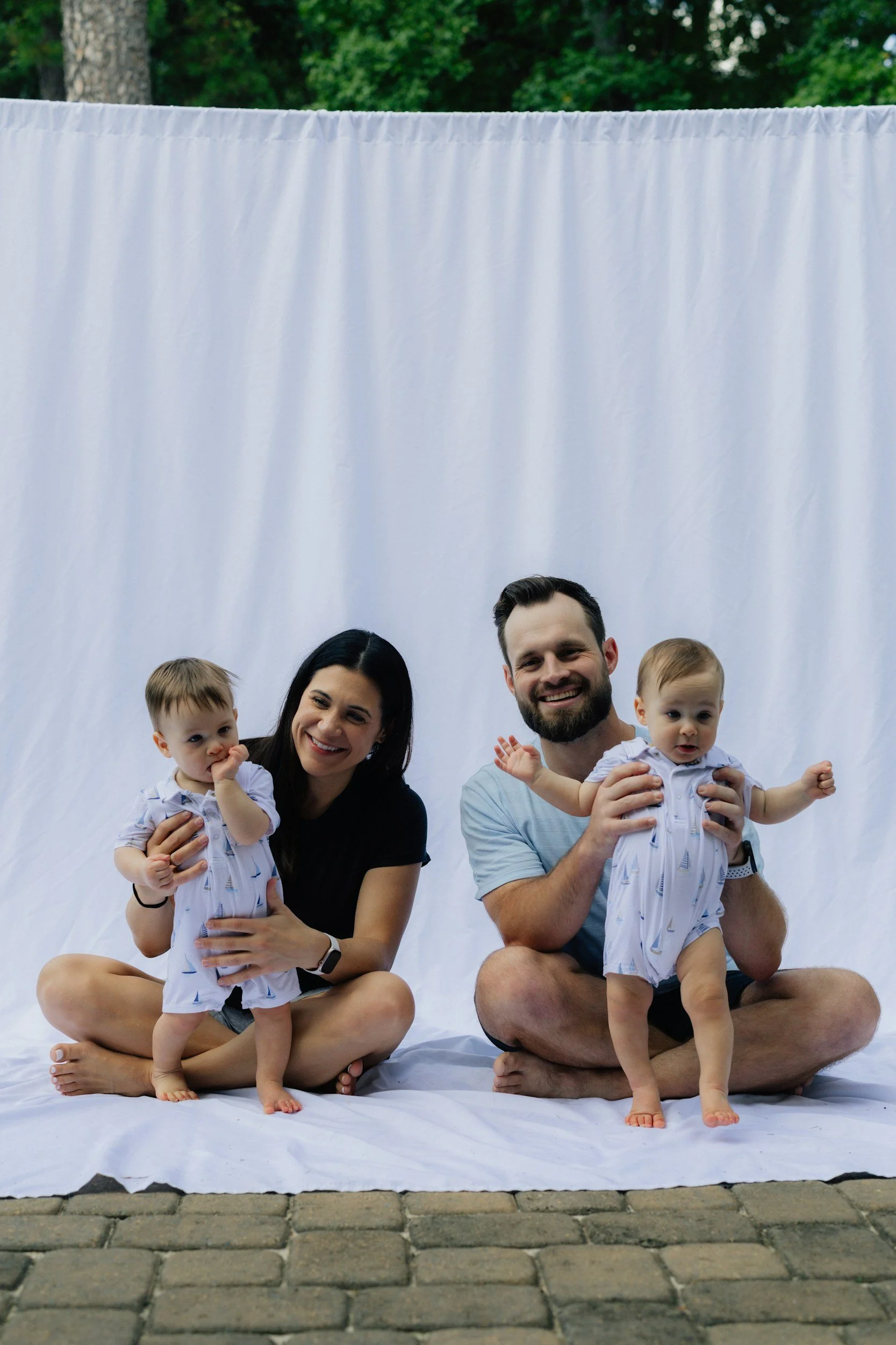 A happy family of four, including a woman, a man, and two toddlers, sitting on a white sheet outdoors with a white cloth backdrop and trees in the background.