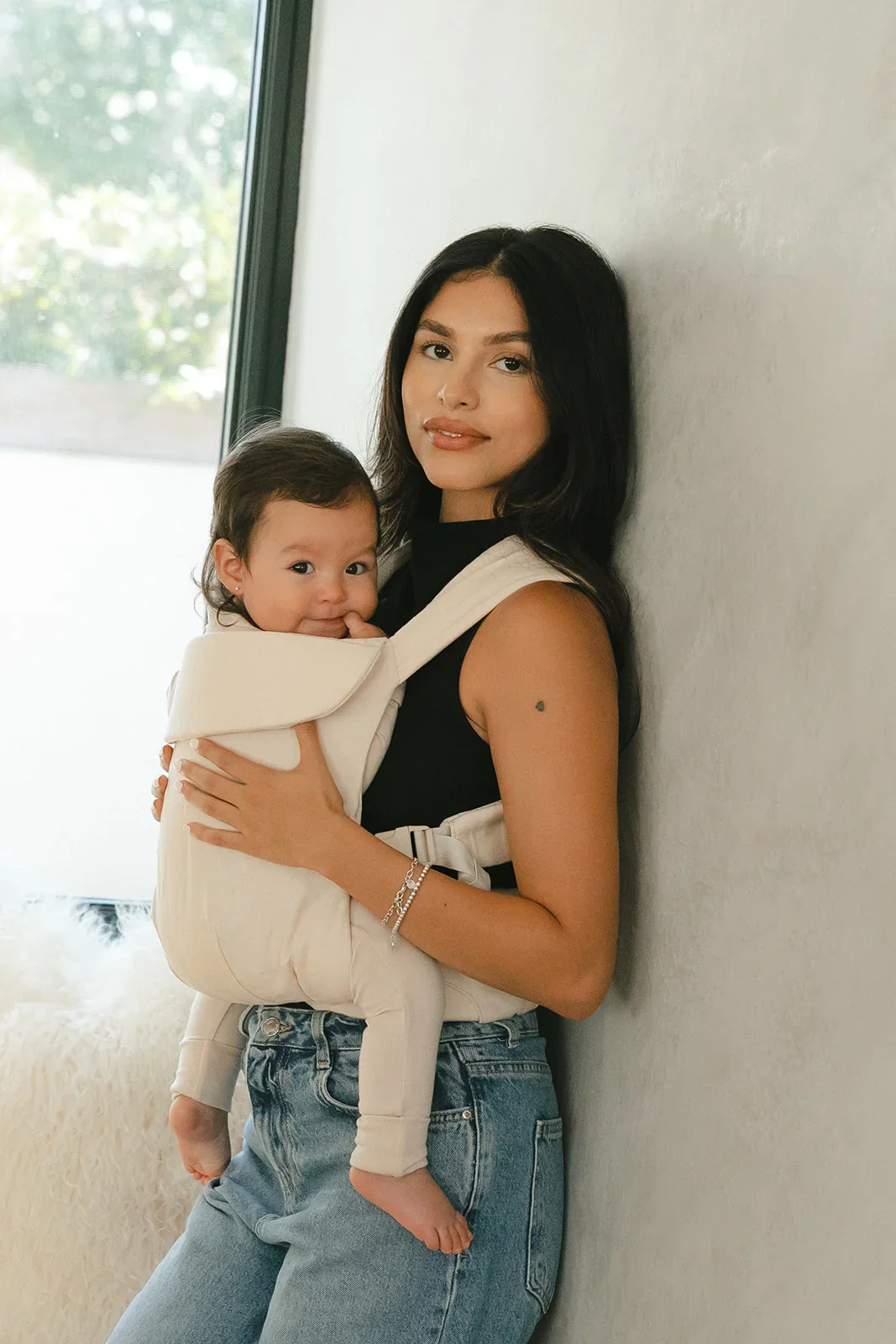A woman with dark hair wearing a black sleeveless top and jeans, holding a young girl in a cream-colored baby carrier. The girl has curly hair and is looking at the camera, resting her hand on her mouth. They are standing indoors near a window.