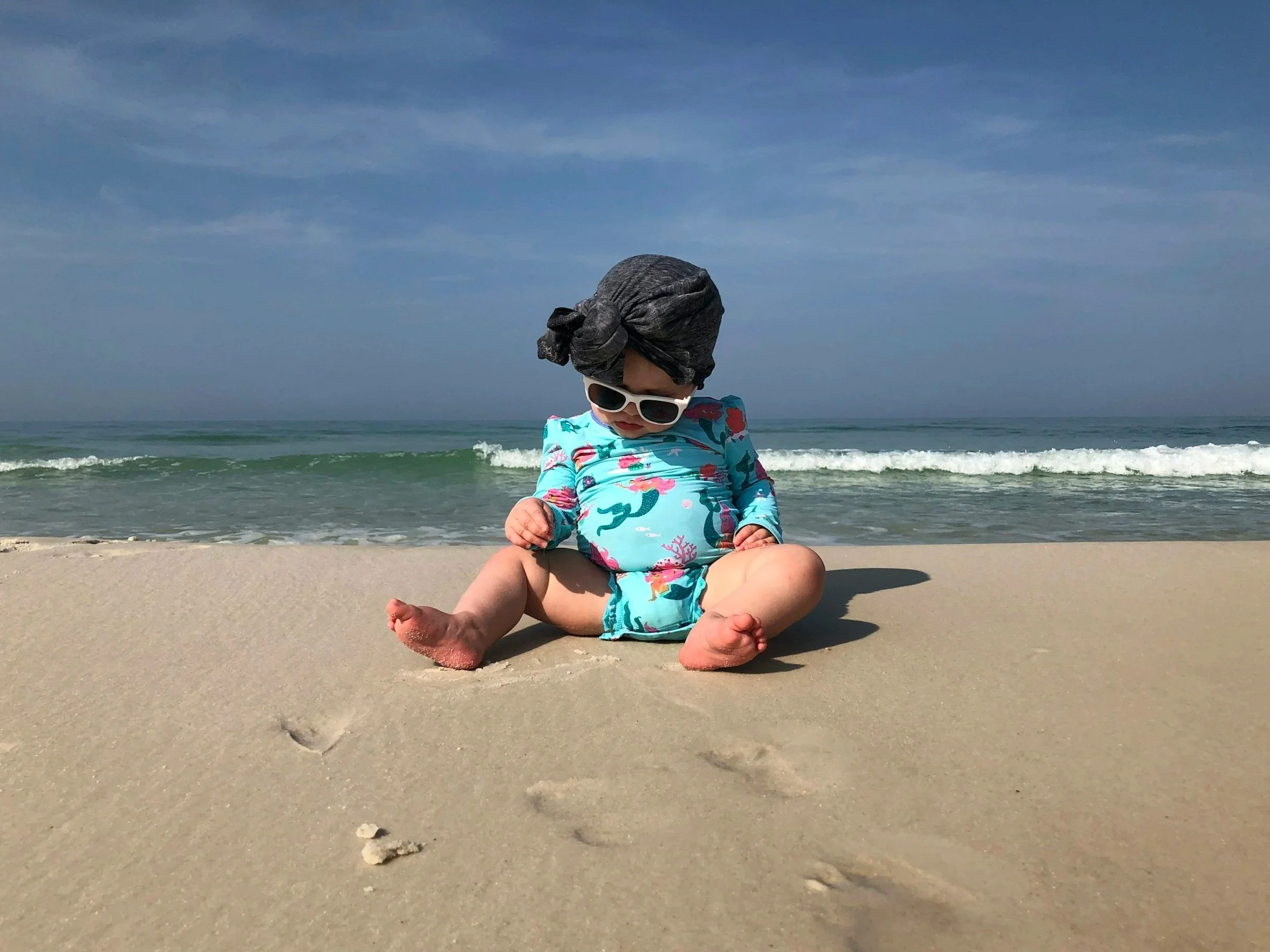 A young child sitting on the sandy beach near the water, wearing a blue swimsuit with pink and green patterns, a black headwrap, and white sunglasses, with ocean waves in the background.