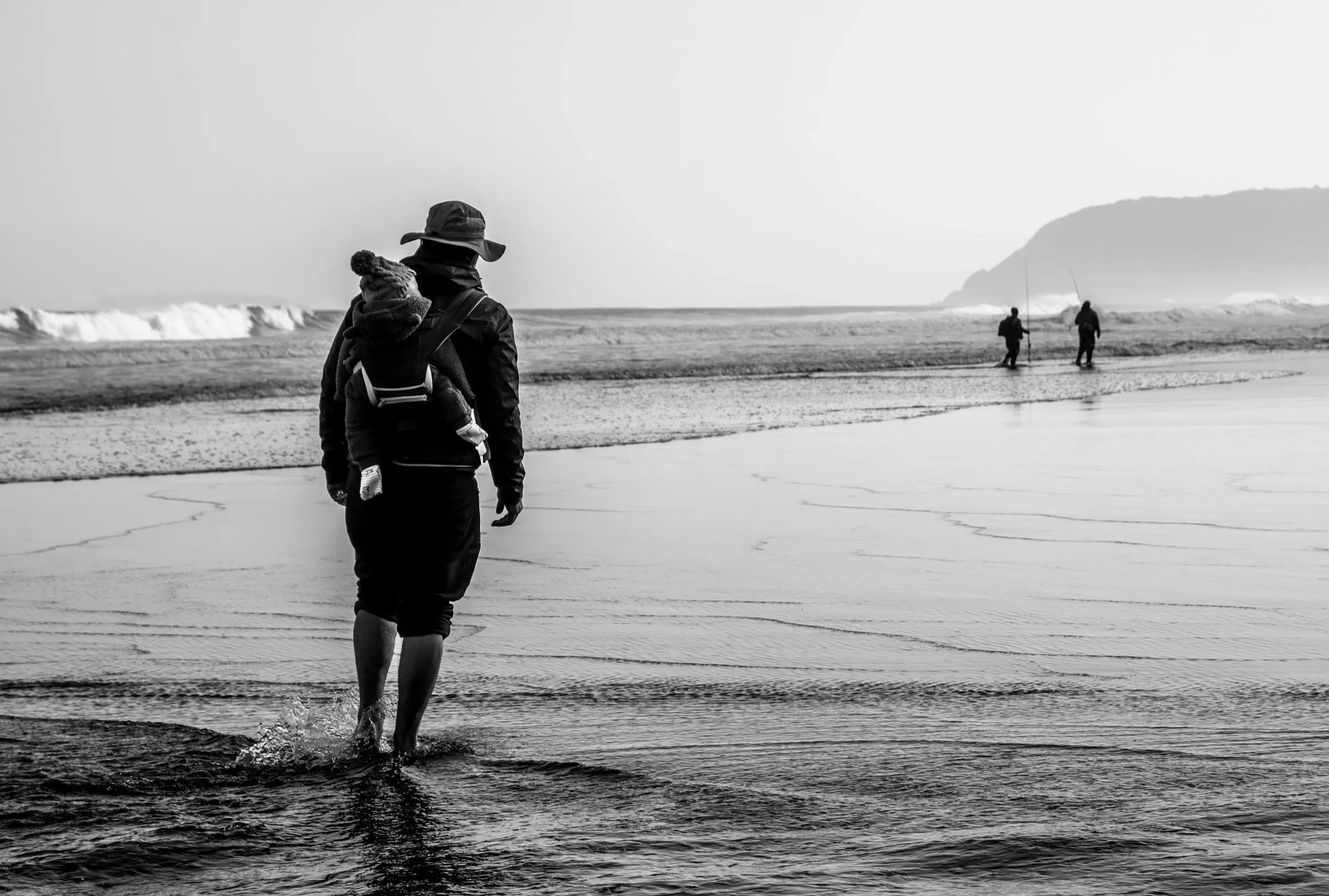 A person with a child in a backpack walking in shallow water at the beach, with two people fishing in the background near the shoreline.