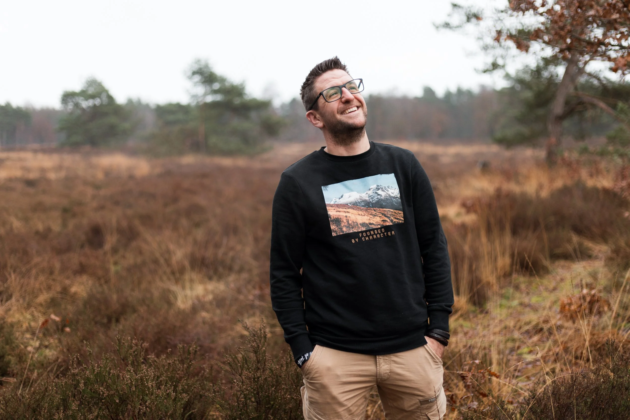 Een lachende man met korte haar en bril, draagt een zwart sweatshirt met een bergafbeelding en de tekst 'Founded by character', staat in een buitenlandschap met herfstbomen en gras.