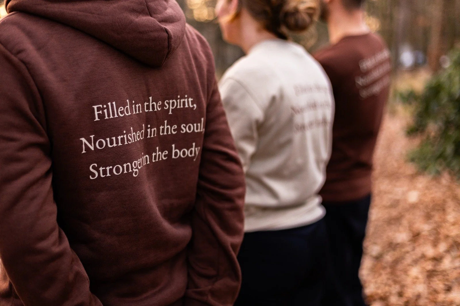 Driedaagse staat er op drie mensen in een bos, waarvan de nadruk ligt op iemand in een bruin hoodie met quote op de achterkant, die zegt 'Filled in the spirit, Nourished in the soul, Strengthen in the body'.