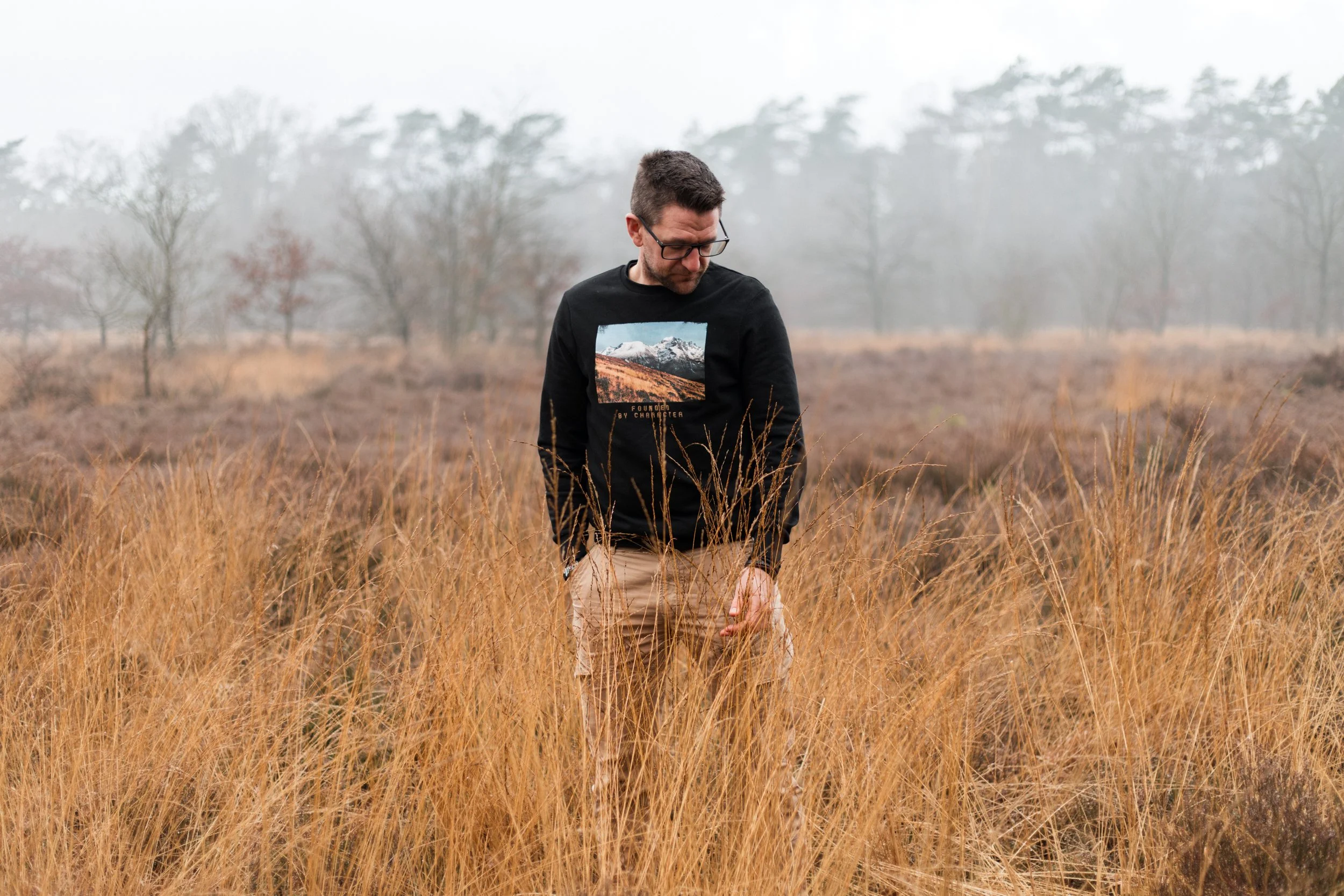 Man staat in een veld met bruin gras, op de achtergrond staan bomen en een mistige hemel.
