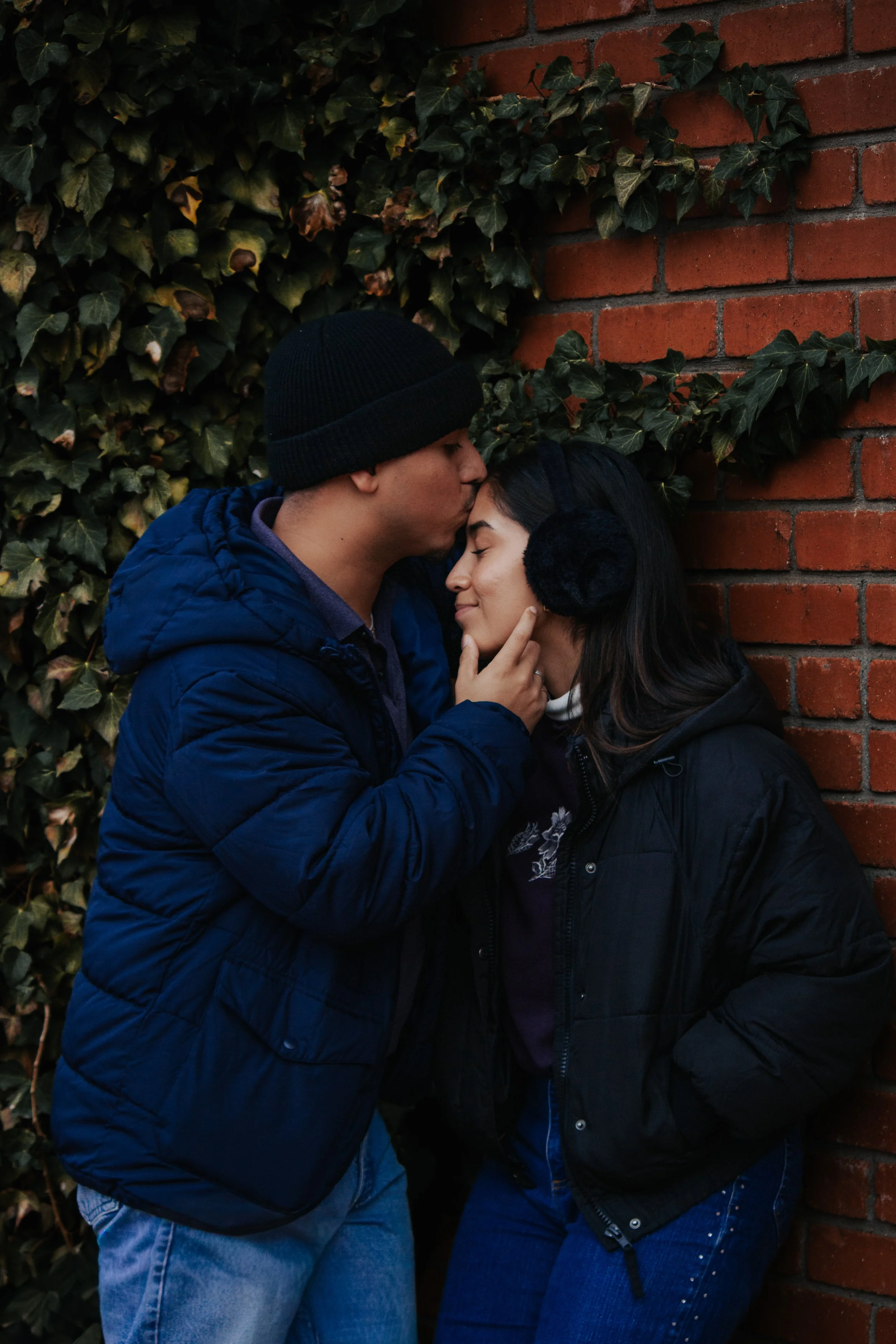 A couple sharing an intimate moment outdoors against a brick wall and green ivy, with the man gently kissing the woman's forehead and both appearing happy and content.