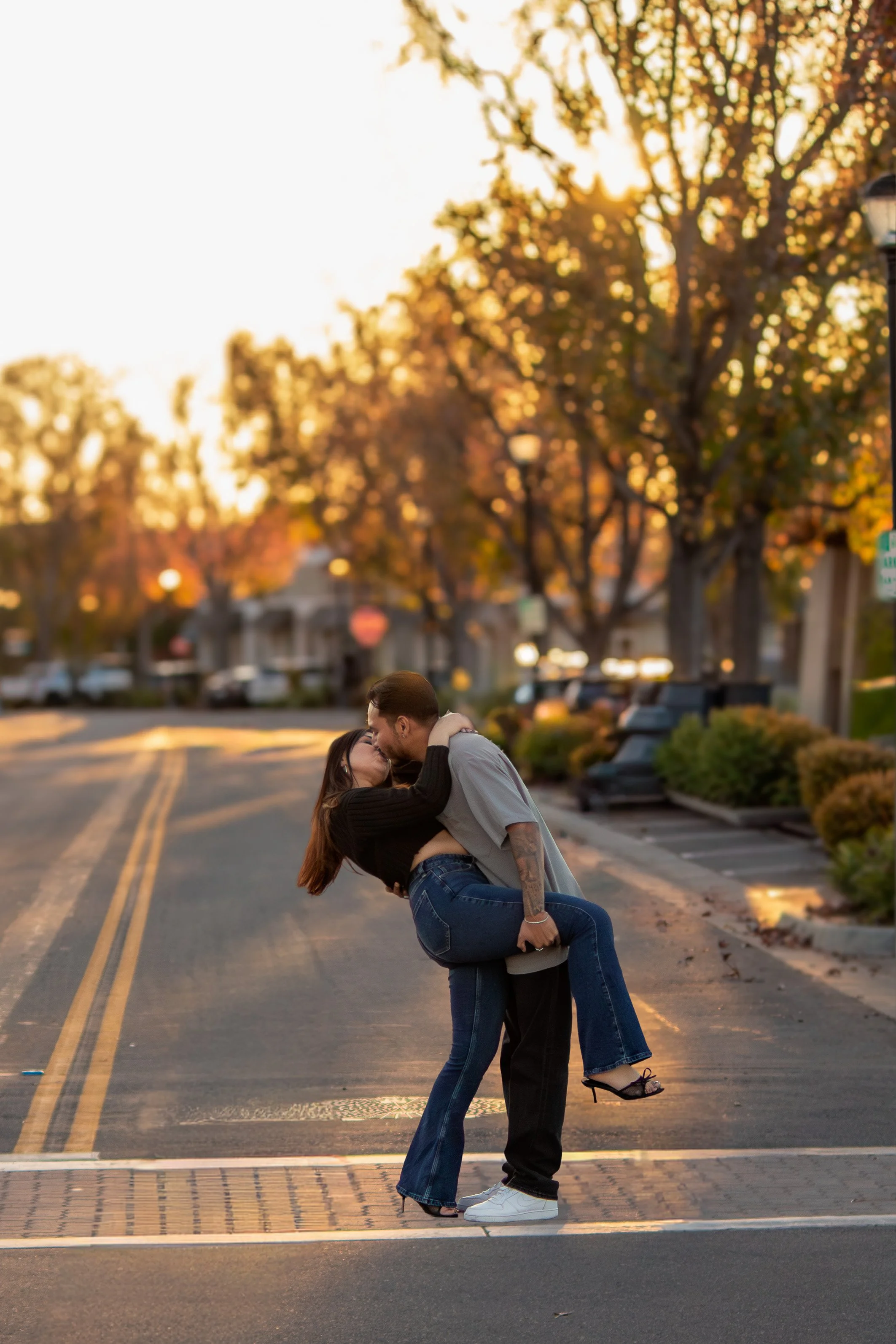 A couple wearing casual clothes dancing and kissing in a street at sunset, with trees and parked cars in the background.
