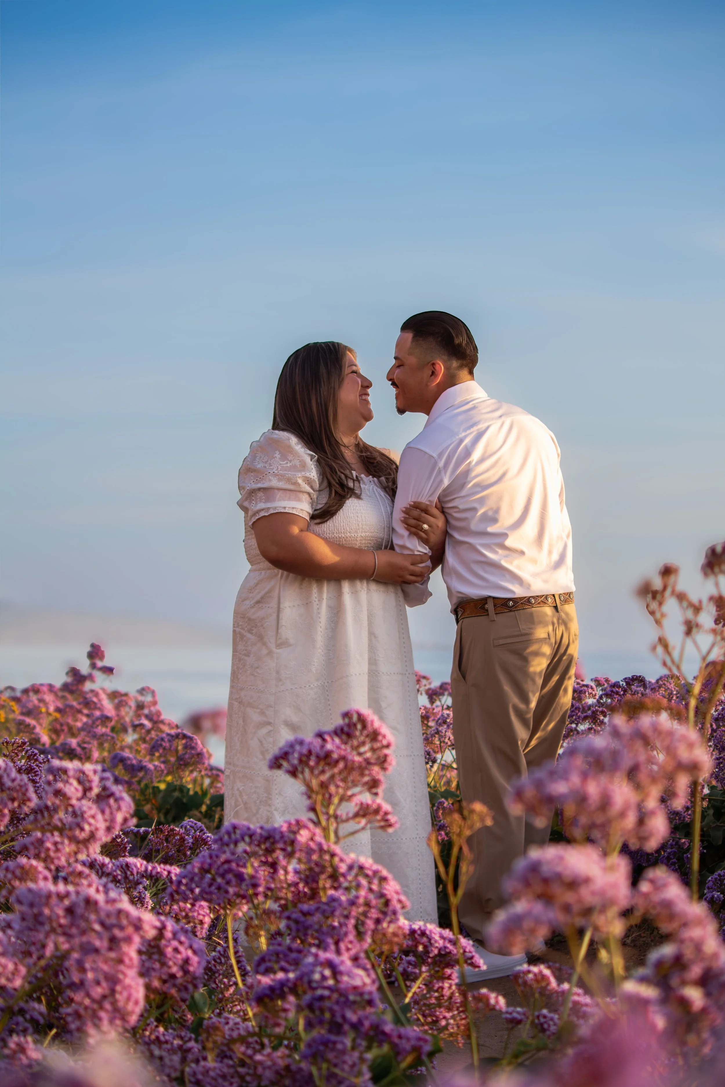 A couple stands in a field of pink and purple flowers, facing each other and smiling, with a blue sky above
