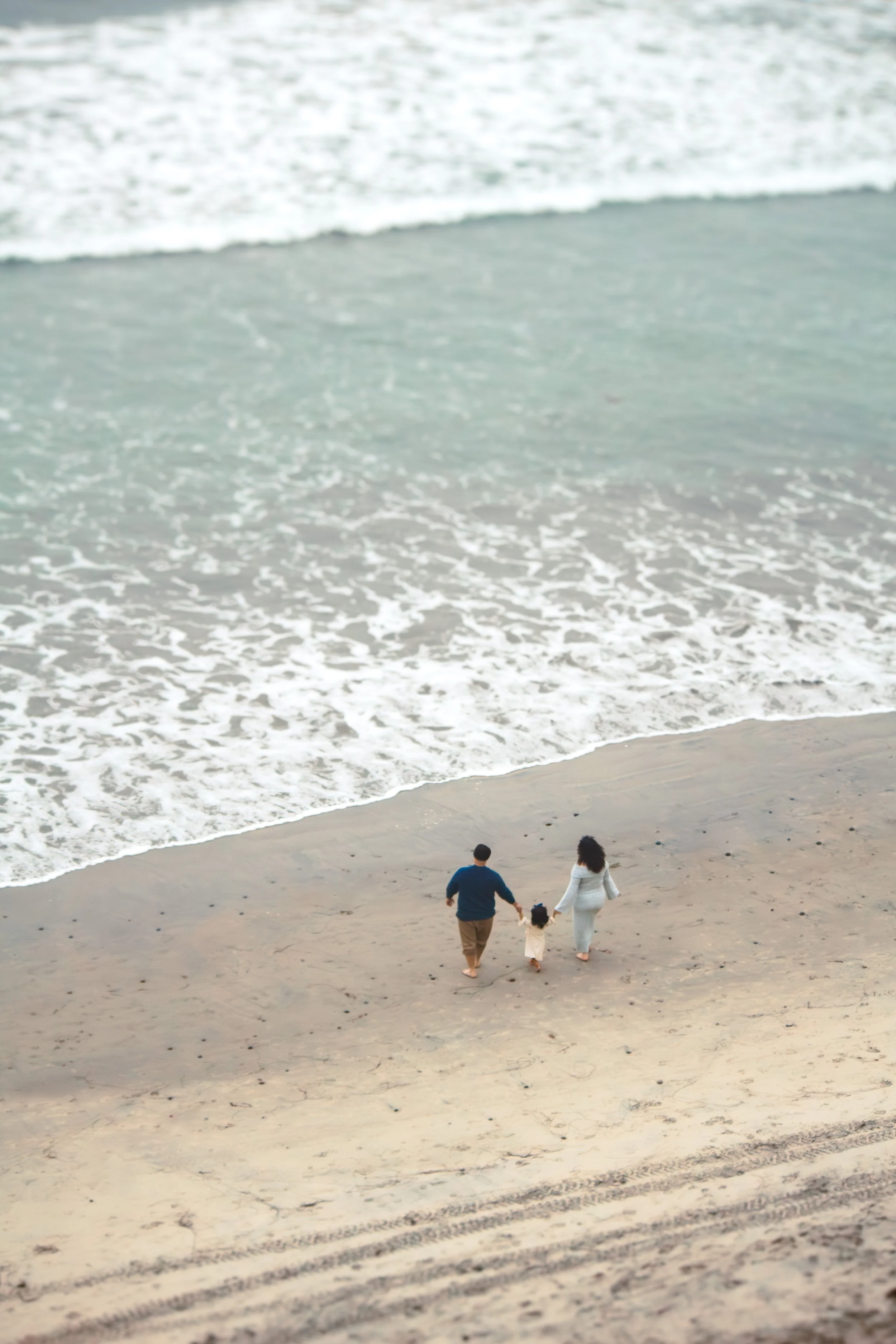 A family of three, two adults and a child, walking hand-in-hand on the sandy beach near the water's edge, with waves gently crashing nearby.