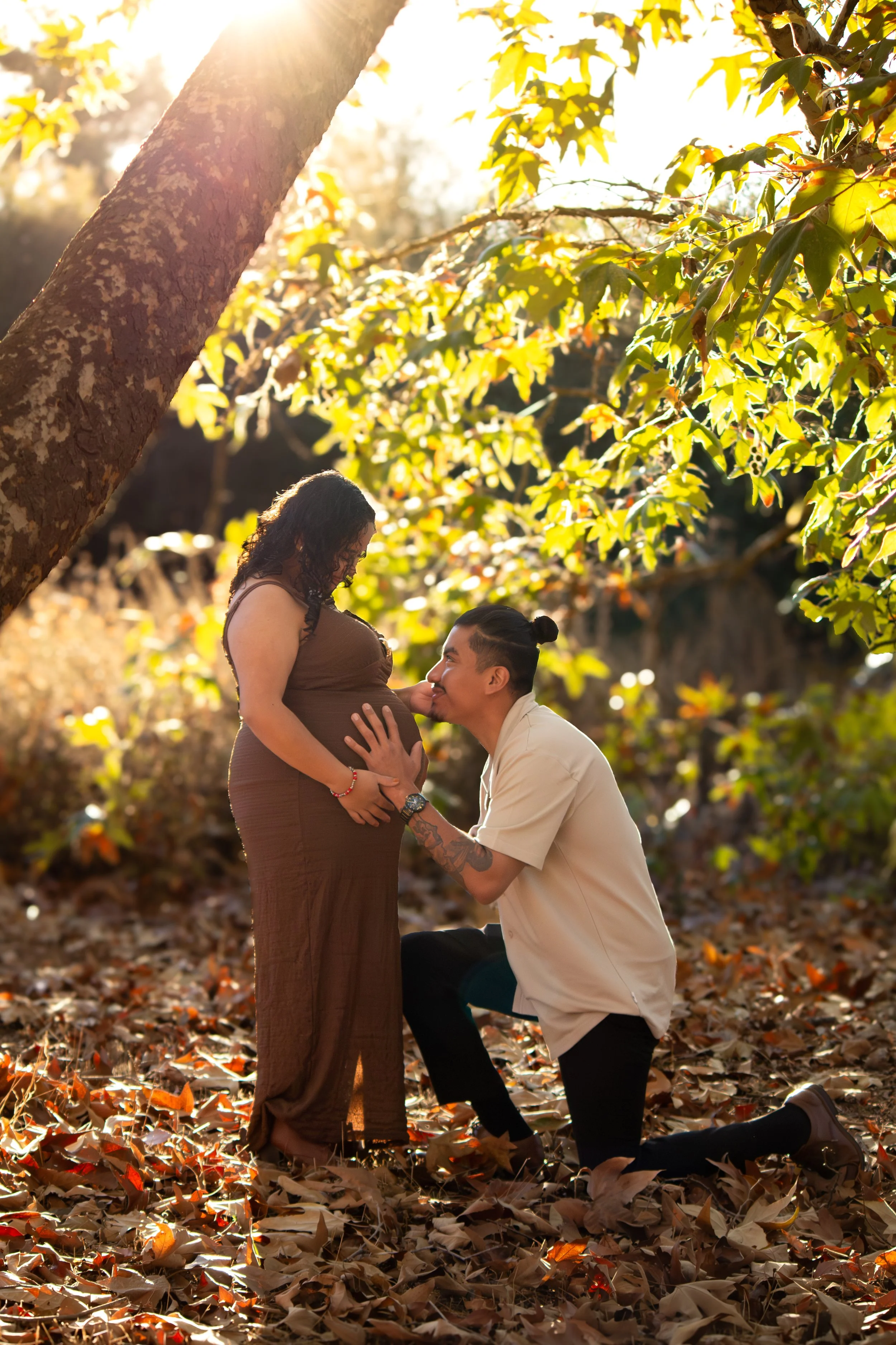 A couple proposing outdoors during autumn, with the woman pregnant, surrounded by fallen leaves and green trees, as the man kneels and holds her hand while she looks down at him.