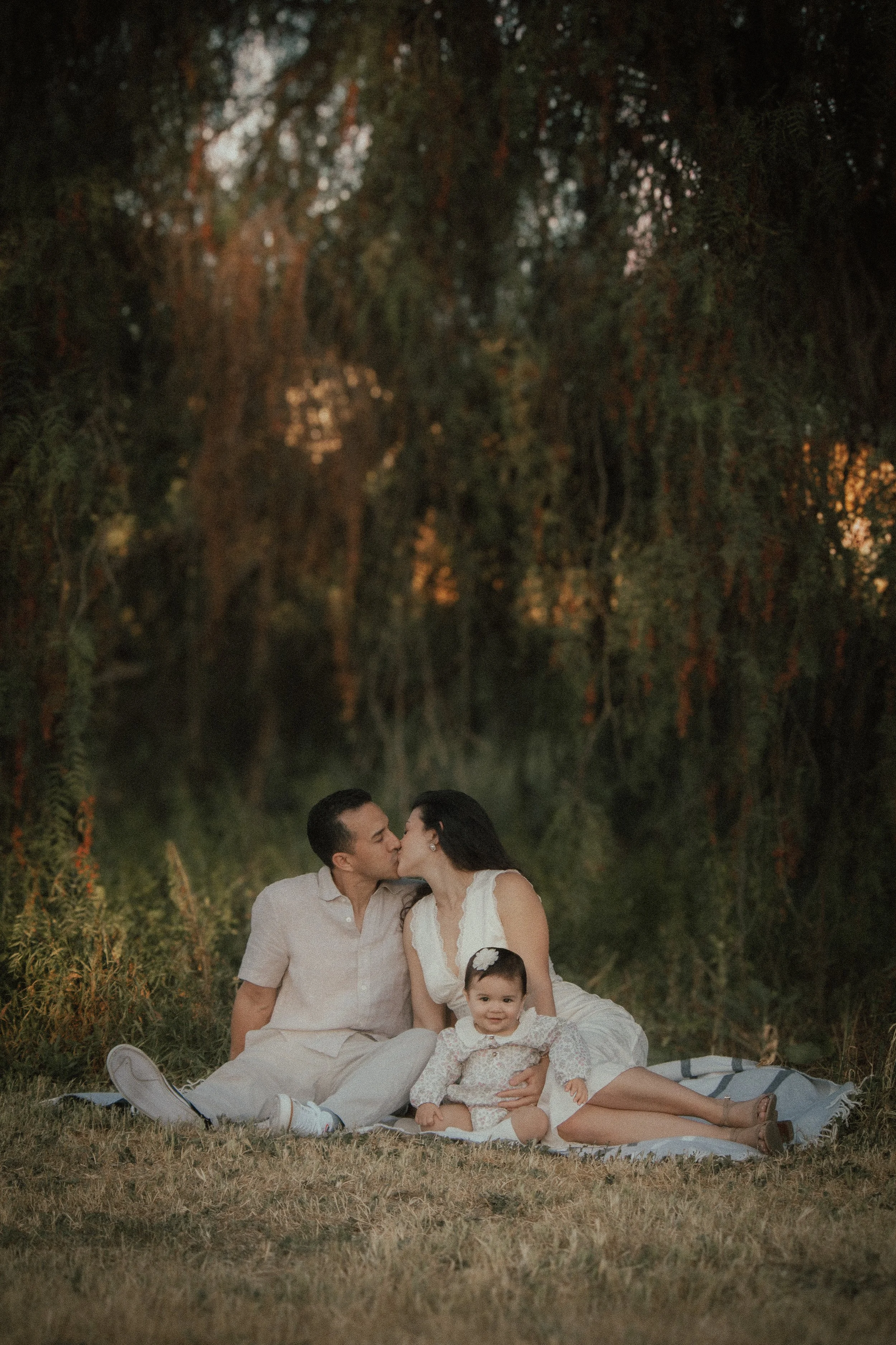 A family of four sitting on a blanket outdoors, with a lush green forest in the background, as they share a kiss.